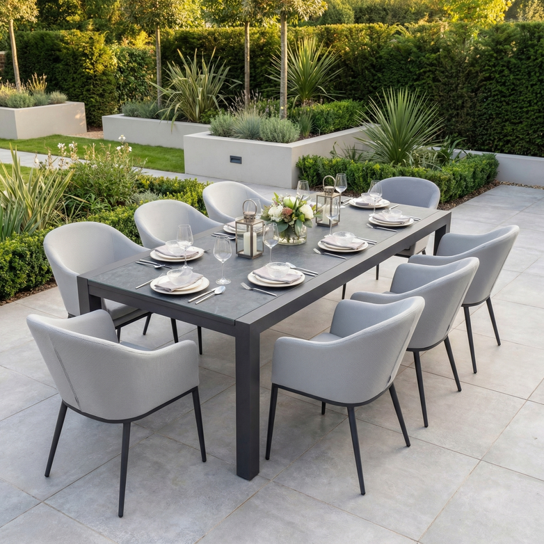 The Luna 8 Seat Outdoor Fabric Extending Ceramic Dining Set in Oyster Grey features stain-proof, stylish comfort with gray chairs, plates, and lanterns—perfect for a landscaped garden gathering.