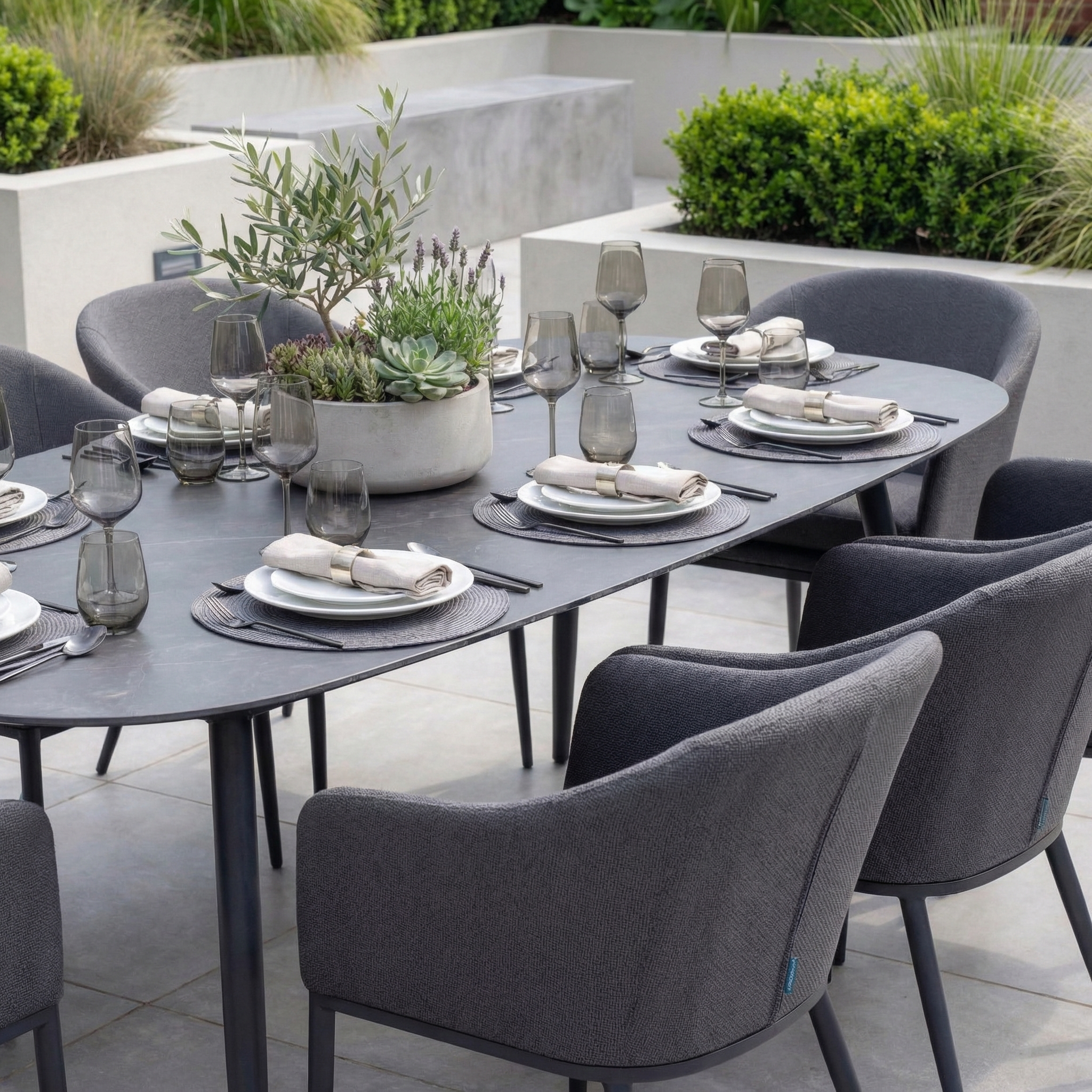 The Luna 8 Seat Outdoor Fabric Oval Ceramic Dining Set in Grey includes stain-resistant chairs and a stylish table, with plates, glasses, and a plant centerpiece arranged for outdoor dining on your patio.