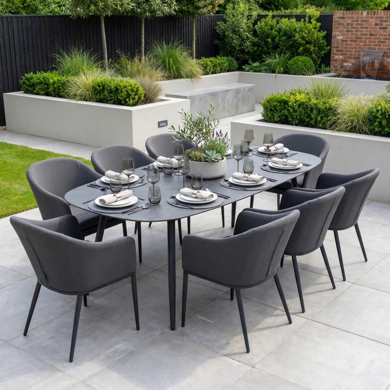 The Luna 8 Seat Outdoor Fabric Oval Ceramic Dining Set in Grey features eight gray chairs and a black table, made from stain-proof materials, elegantly arranged with plates and glasses on a garden patio.