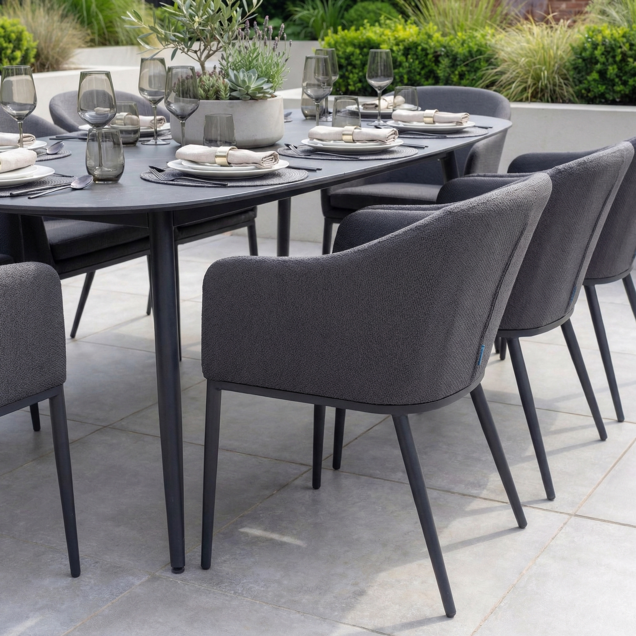 The Luna 8 Seat Outdoor Fabric Oval Ceramic Dining Set in Grey features gray cushioned chairs and a ceramic table, set with glassware and plates on a patio. Crafted from all-weather materials for enduring style.
