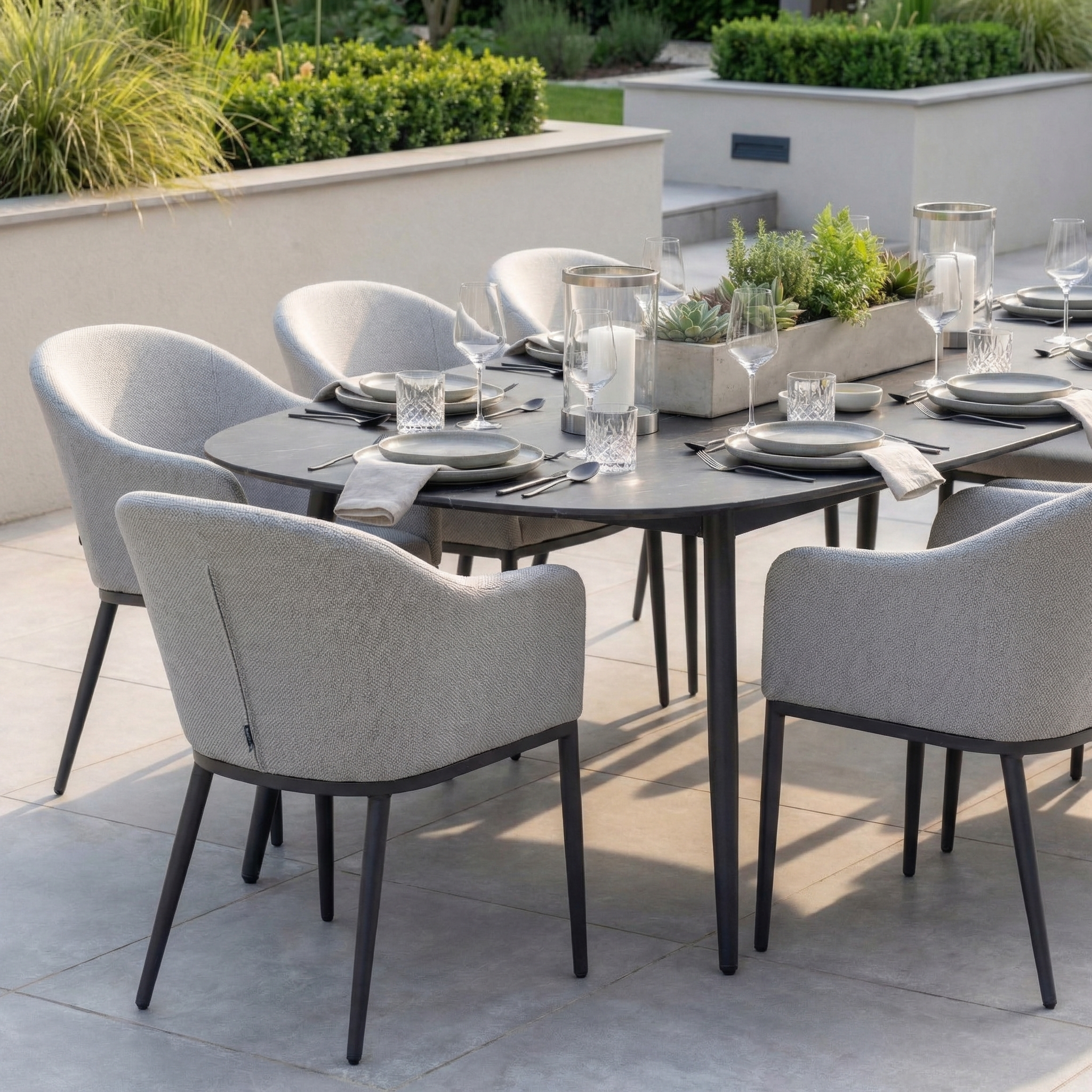 The Luna 8 Seat Outdoor Fabric Oval Ceramic Dining Set in Oyster Grey features gray chairs, plates, glasses, napkins, and a potted plant centerpiece for stylish outdoor dining.