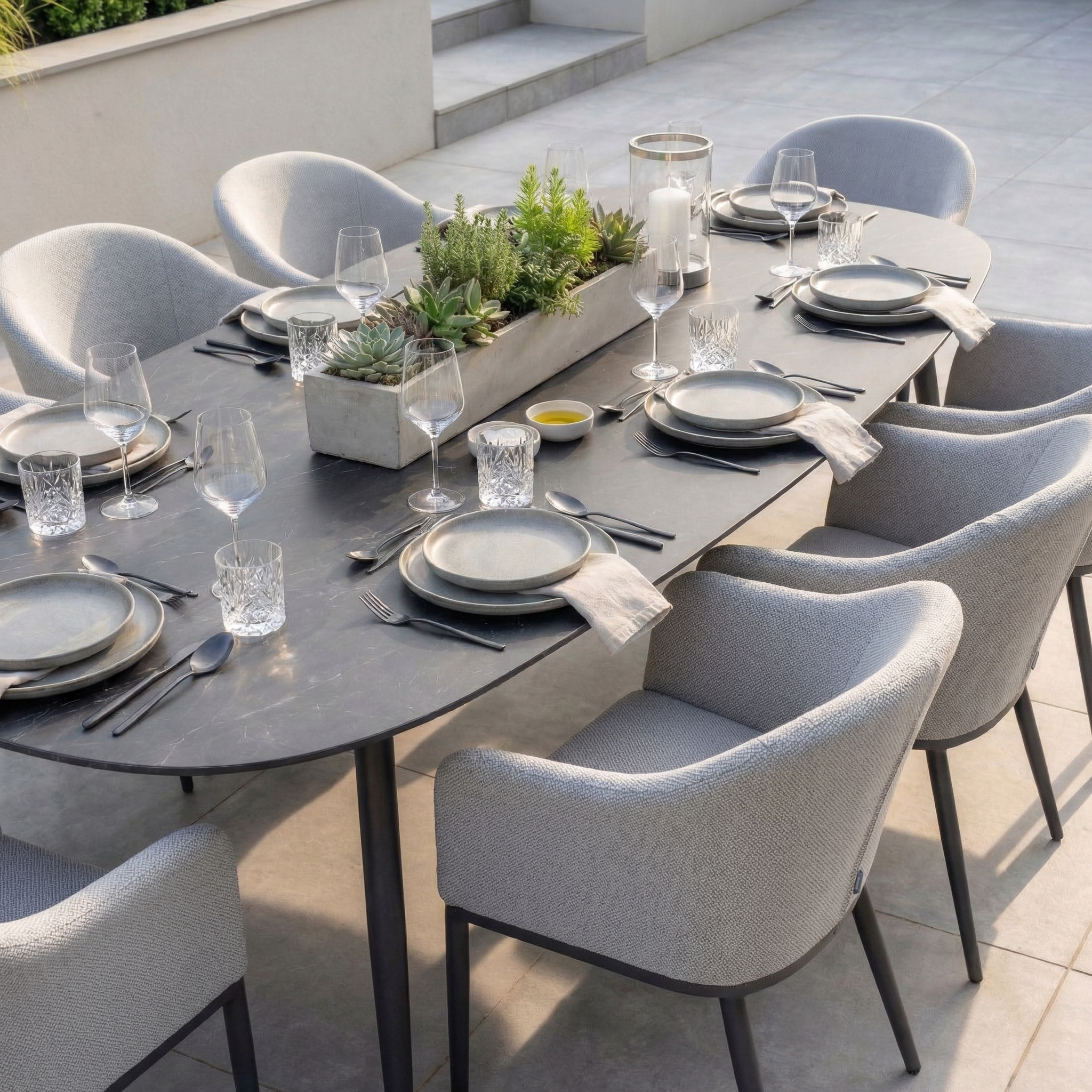 The Luna 8 Seat Outdoor Fabric Oval Ceramic Dining Set in Oyster Grey features modern gray chairs, durable all-weather fabric, glassware, and a succulent centerpiece for elegant outdoor dining.