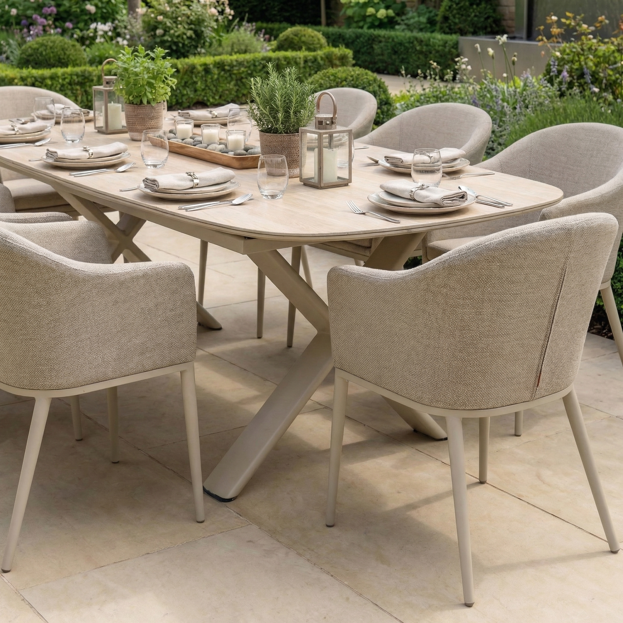 The Luna 8 Seat Outdoor Fabric Oval Dining Set with Albatross Extending Table in Fawn includes beige chairs, plates, glasses, and potted plants arranged on a patio, surrounded by lush greenery.