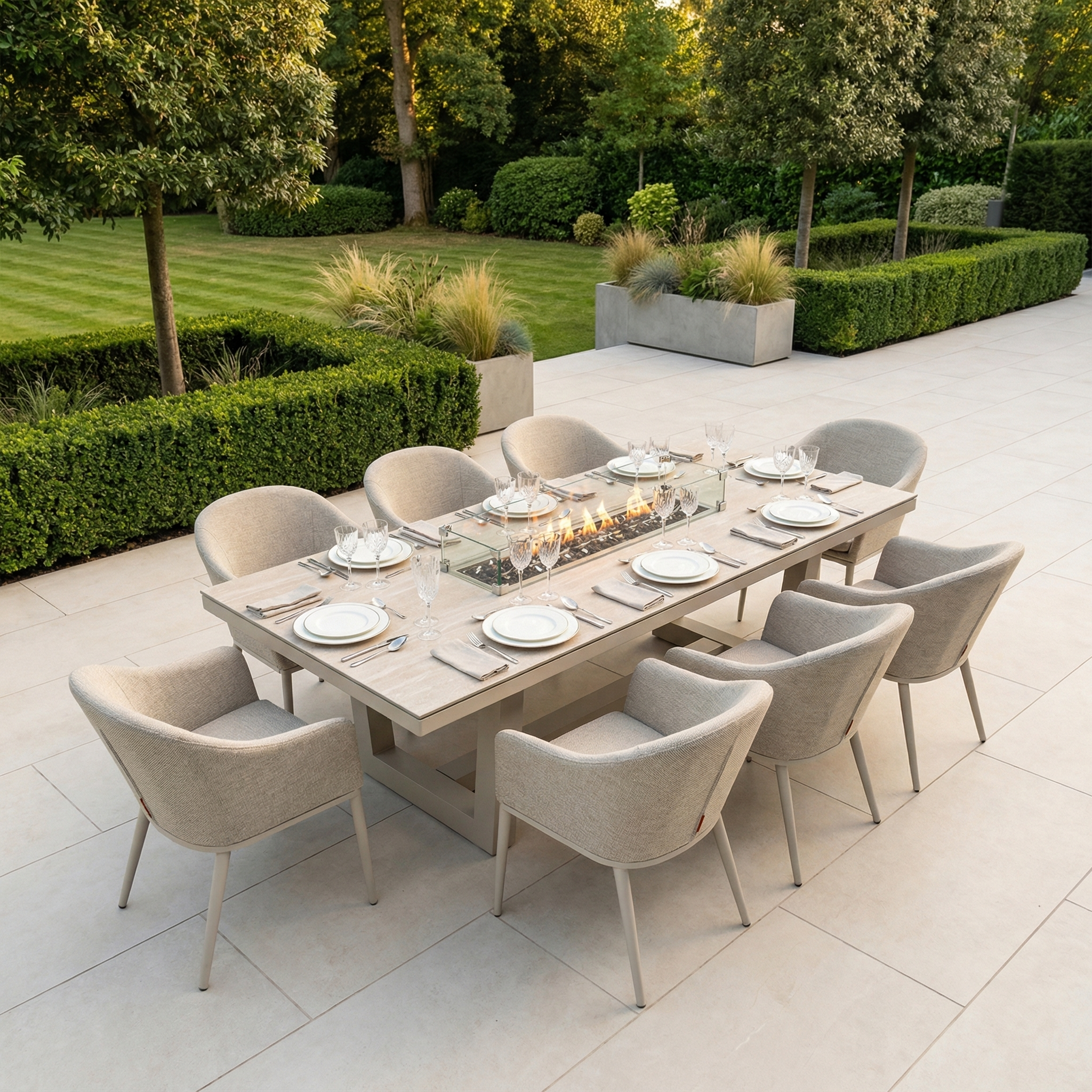 The Luna 8 Seat Outdoor Fabric Rectangular Ceramic Firepit Dining Set in Fawn features a stylish glass firepit, all-weather materials, and comfortable seating for eight—perfect for alfresco dining amid greenery and manicured hedges.