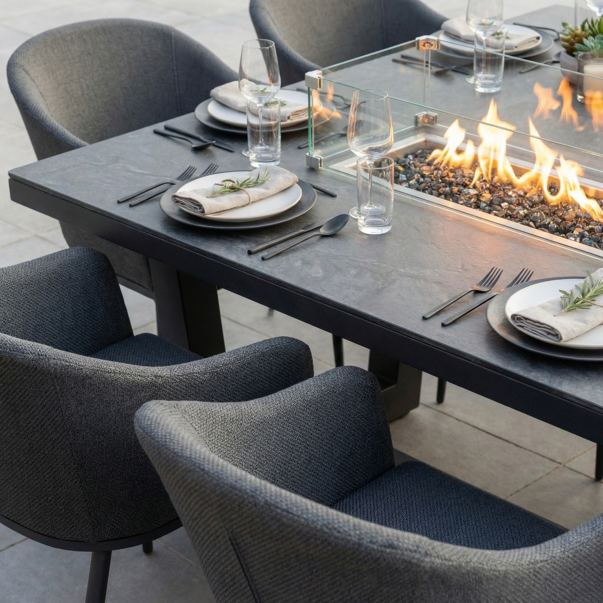 The Luna 8 Seat Outdoor Fabric Rectangular Ceramic Firepit Dining Set in Grey features all-weather grey chairs and a central glass fire pit, set with plates and glasses for stylish outdoor dining.