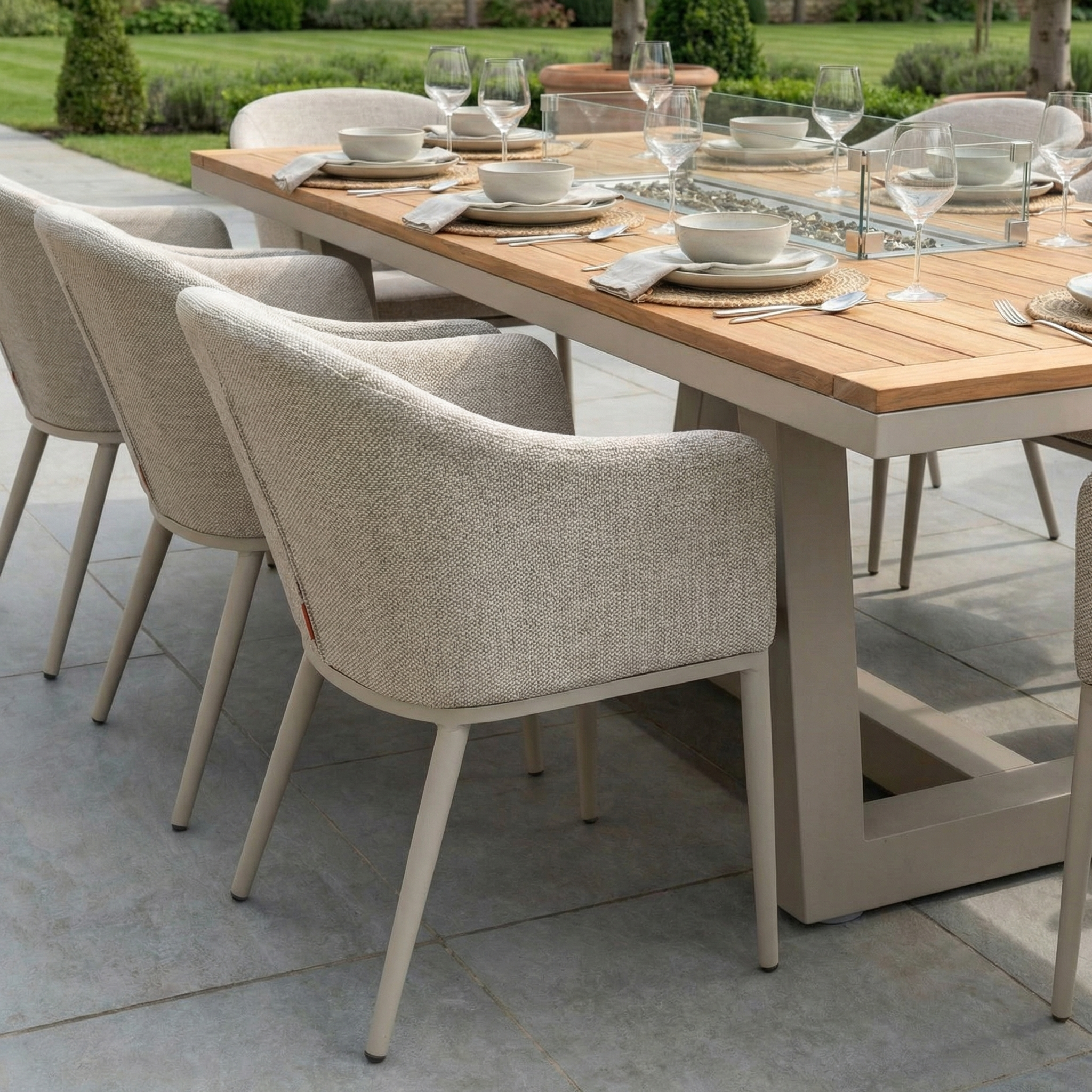 The Luna 8 Seat Outdoor Fabric Rectangular Teak Firepit Dining Set in Fawn is arranged for dining on a patio, offering all-weather comfort and views of the garden.
