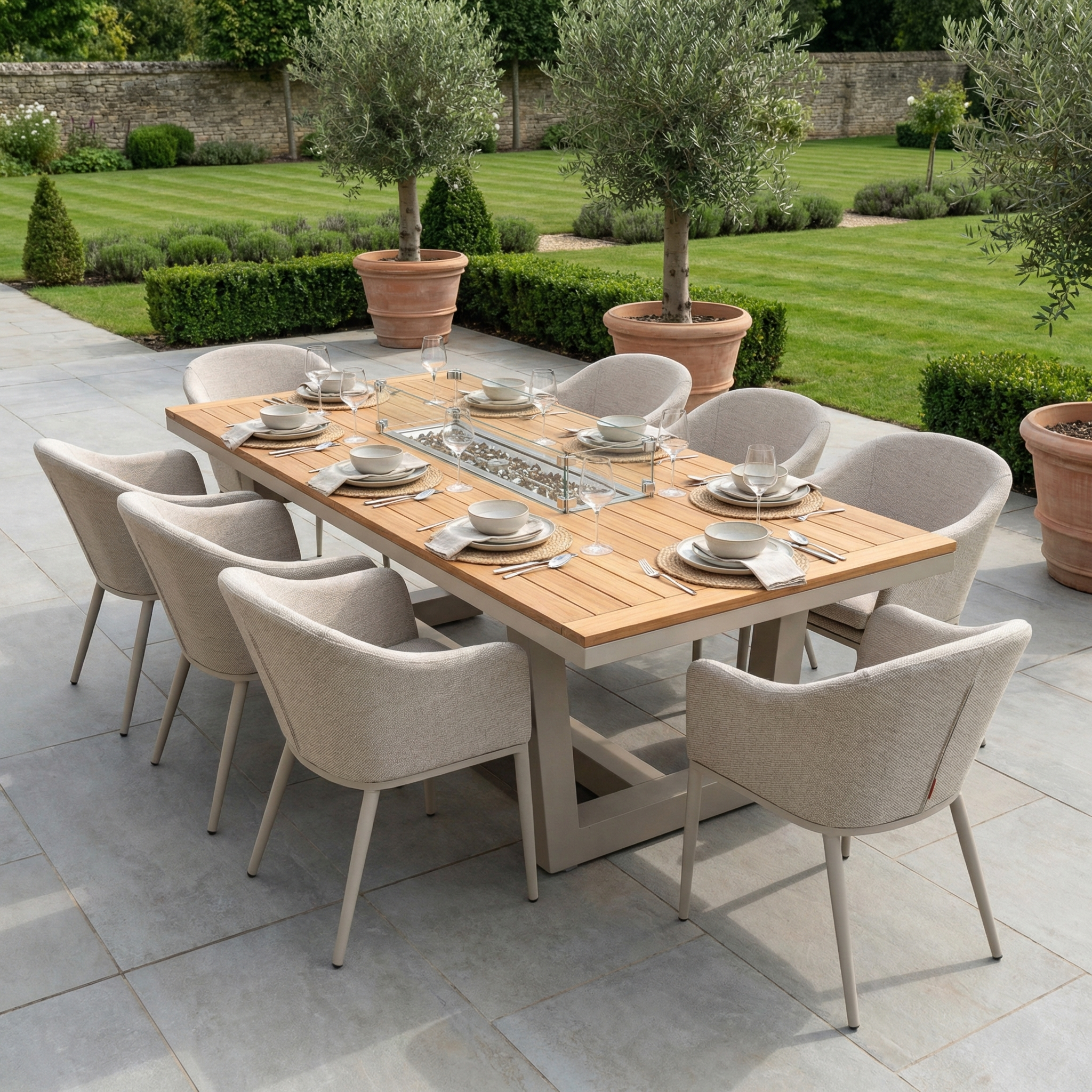 The Luna 8 Seat Outdoor Fabric Rectangular Teak Firepit Dining Set in Fawn is arranged on a patio for eight, with lush greenery and trees providing a scenic backdrop.