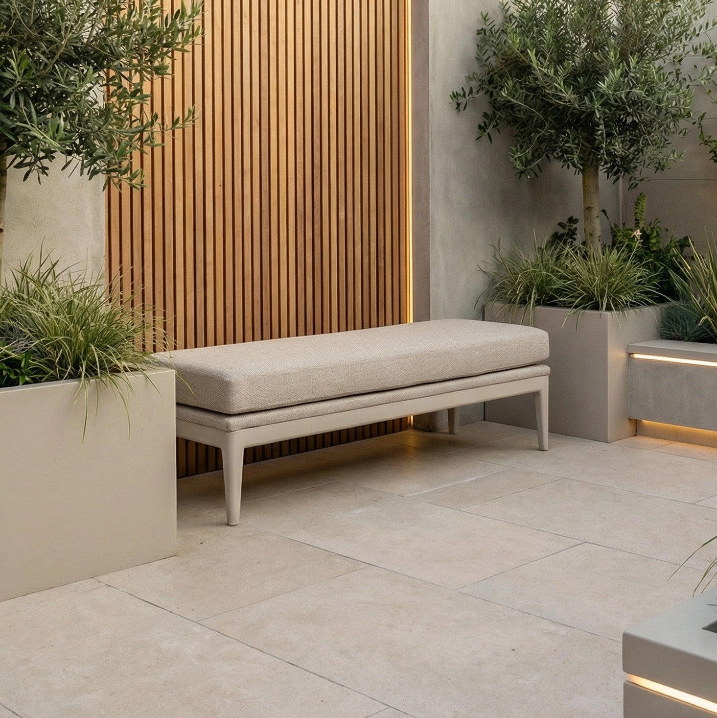 A Luna Bench in Fawn provides outdoor seating between potted plants on a tiled patio, with a wooden slat wall as the backdrop.