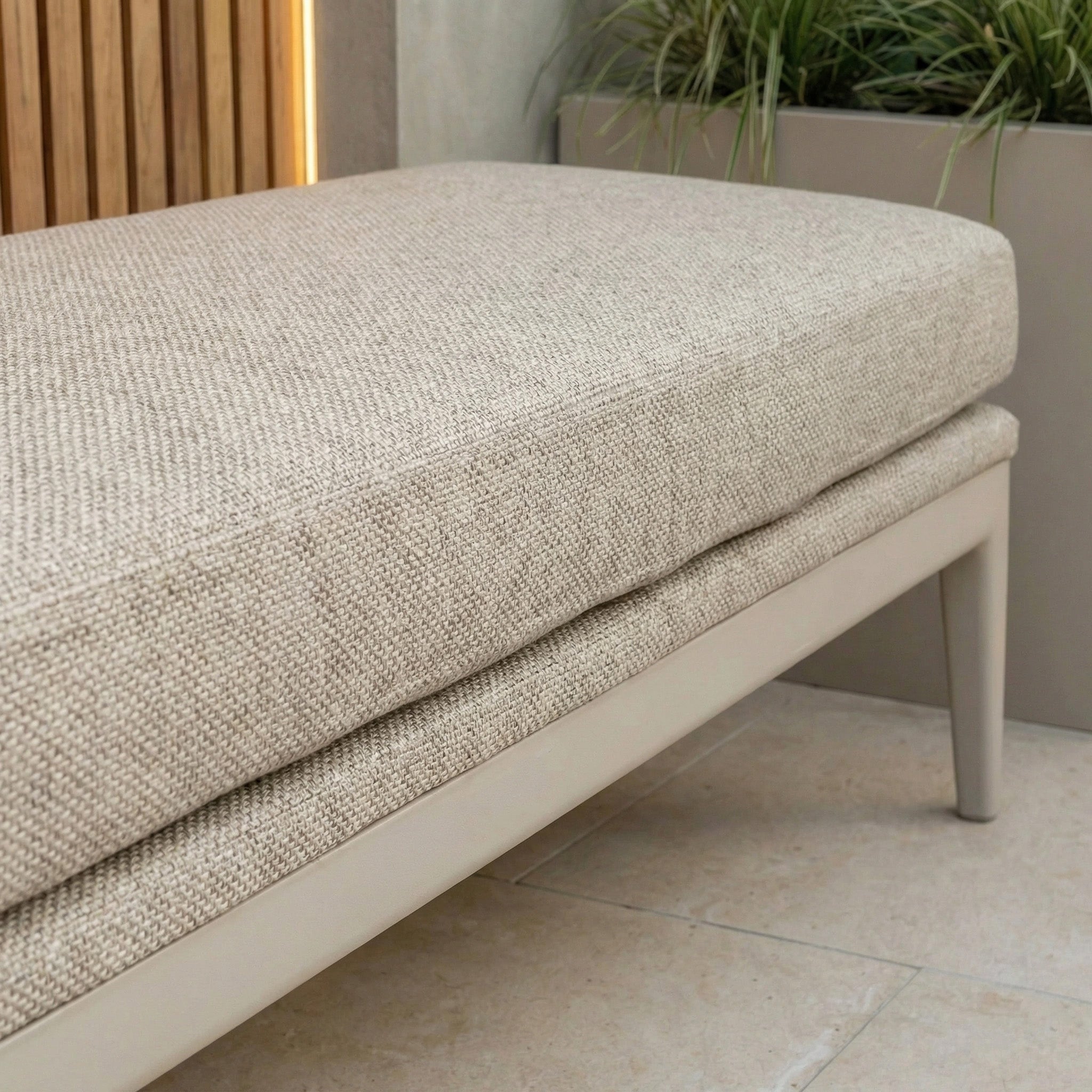 Close-up of the Luna Bench in Fawn, an all-weather bench with a beige cushioned seat and light wood legs, shown beside a planter with green grass—perfect for stylish outdoor seating.
