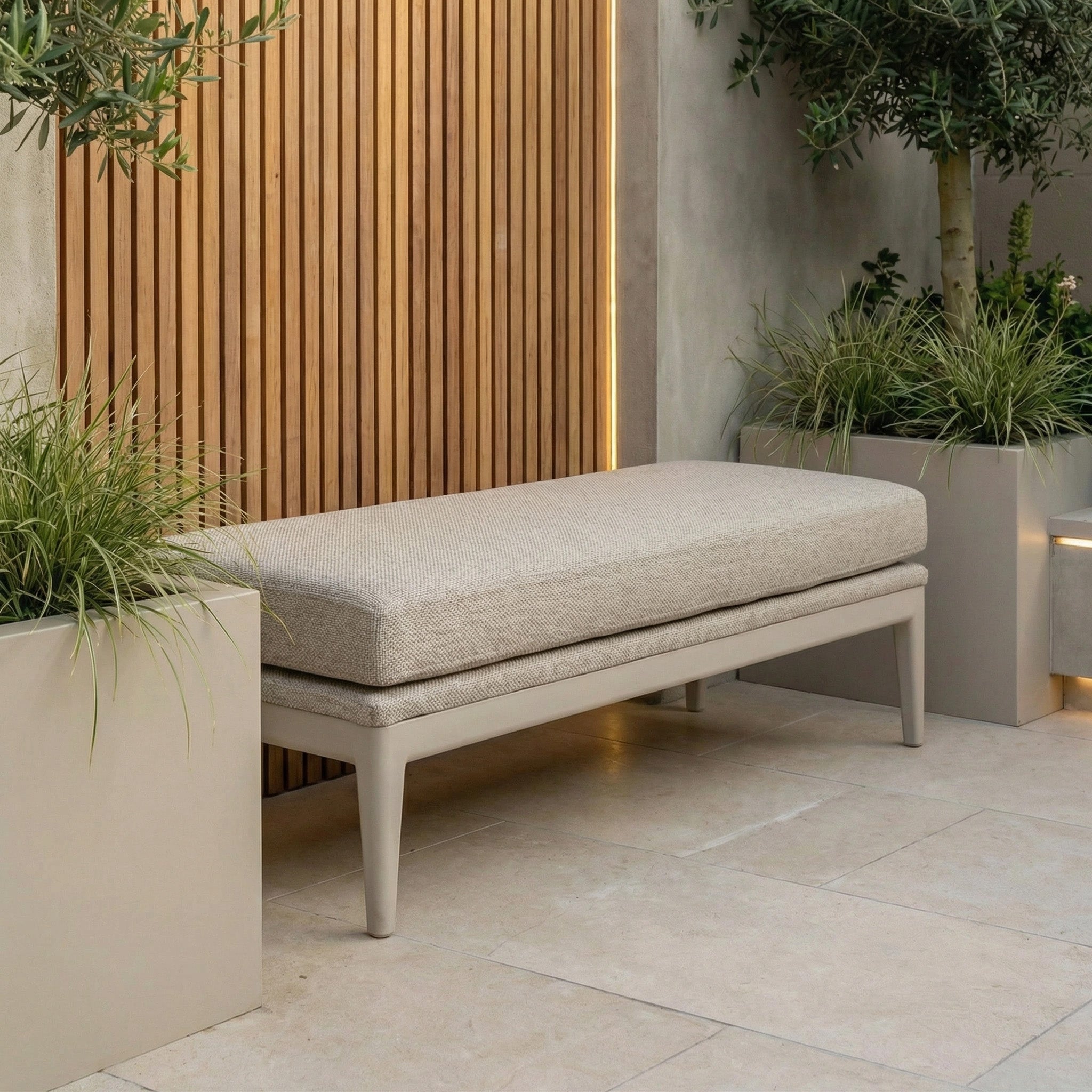 The Luna Bench in Fawn, designed for all-weather use with a cushion, is placed beside tall planters and a wooden slat wall in this modern outdoor space.