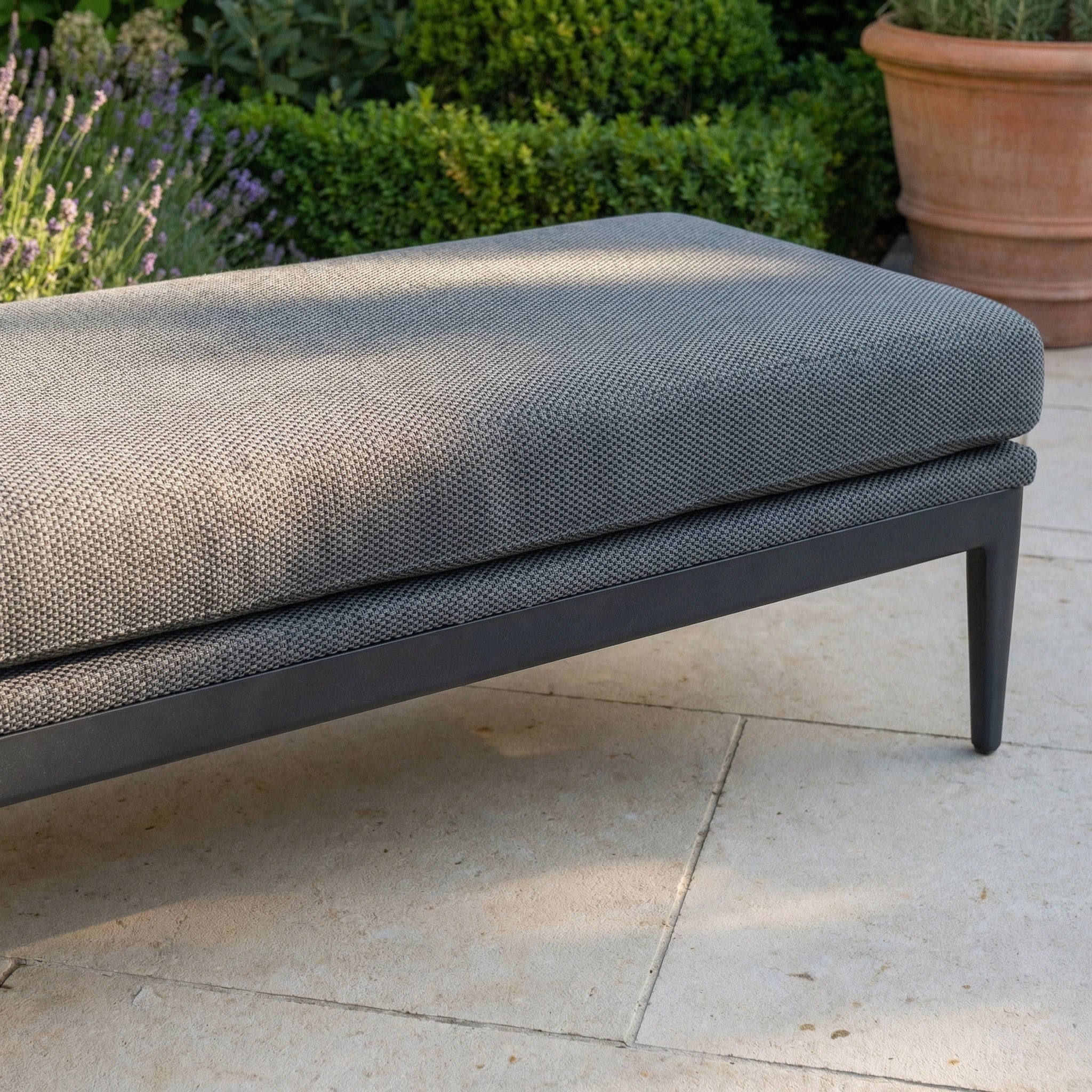 The Luna Bench in Grey, designed for all-weather outdoor use with a textured cushion, is placed on a stone patio amidst lush greenery.