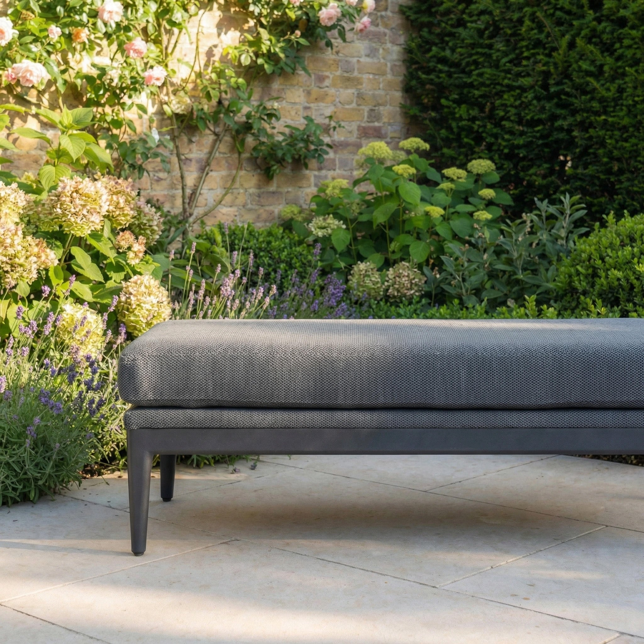 The Luna Bench in Grey is placed on a stone patio amidst flowers and lush greenery.