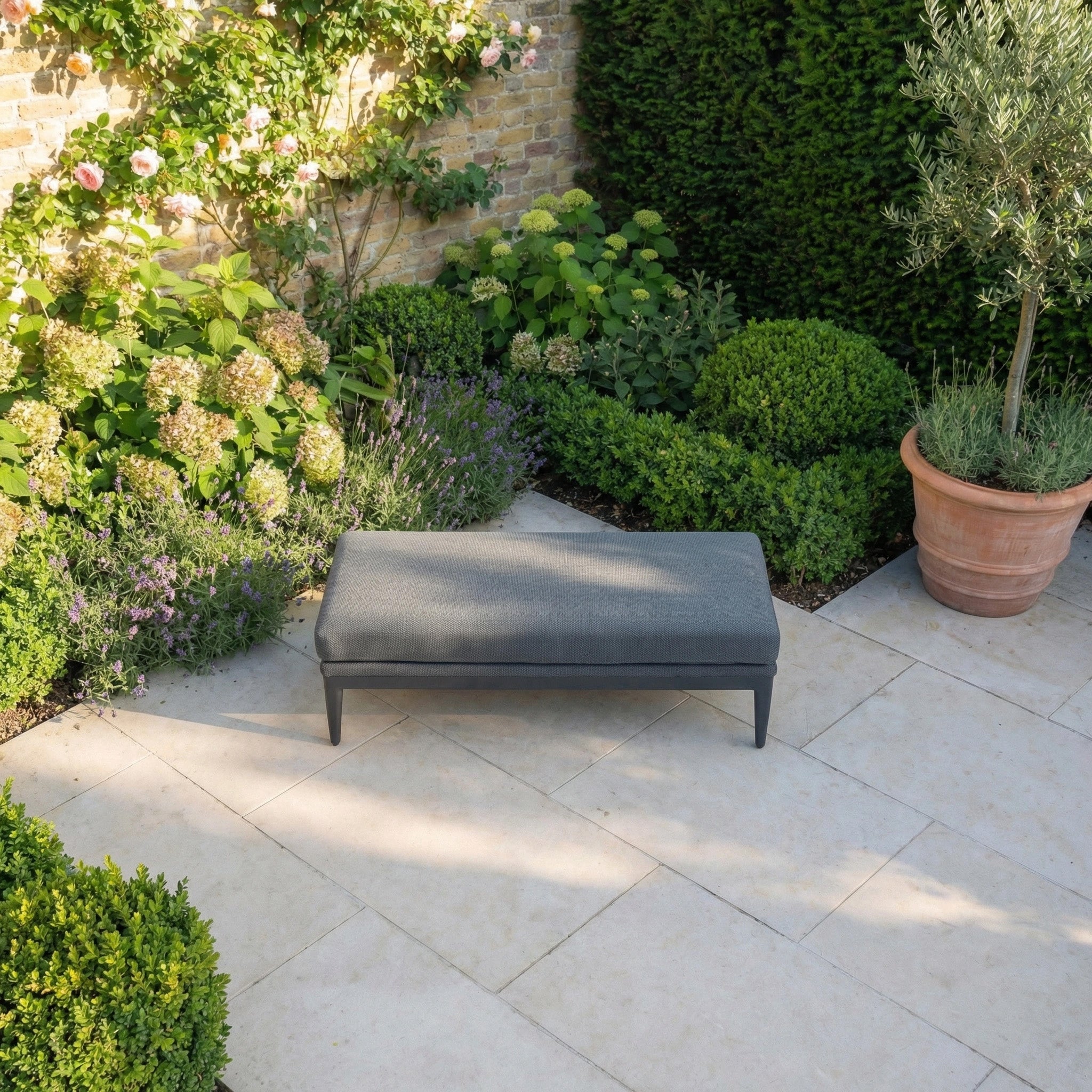 The Luna Bench in Grey, an all-weather bench, rests on a stone patio bordered by green bushes, blooming plants, and a potted tree.