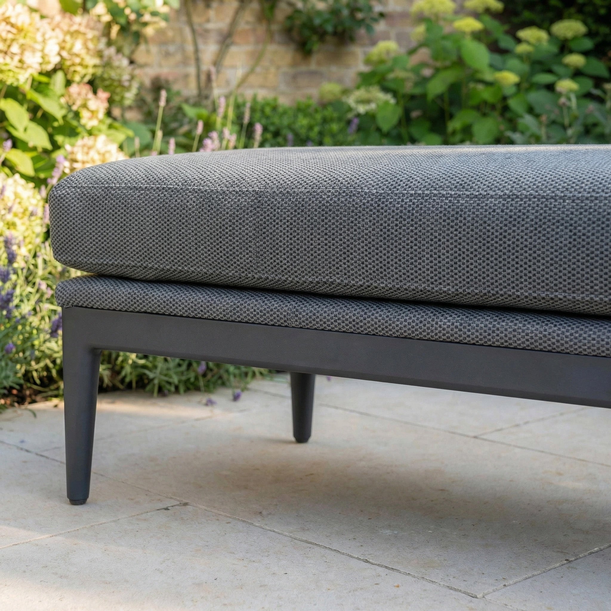 Close-up of the Luna Bench in Grey on a patio, with green plants and a brick wall in the background.