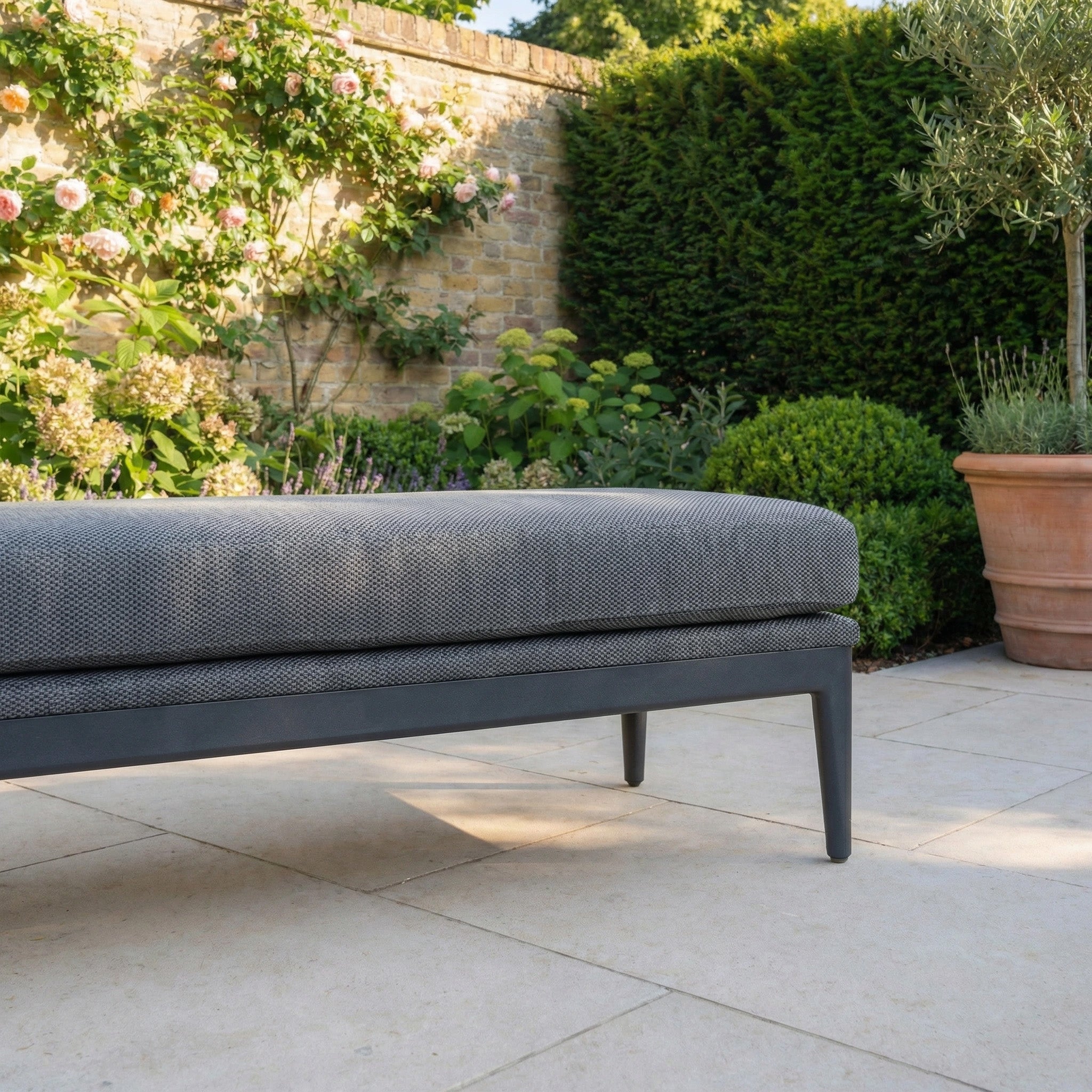 The Luna Bench in Grey, an all-weather outdoor bench, sits on a stone patio amid blooming flowers, lush green bushes, and a classic brick garden wall.