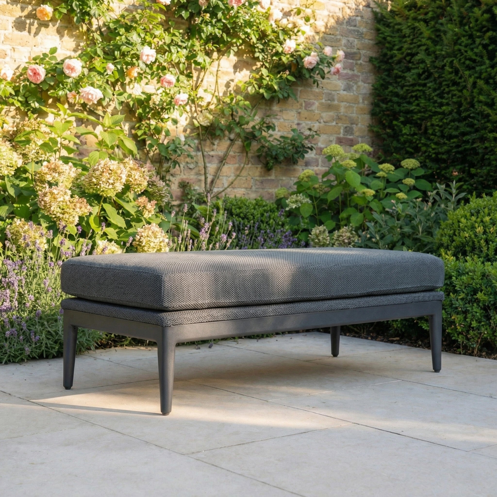 The Luna Bench in Grey is an elegant outdoor seat placed on a stone patio, surrounded by blooming flowers and lush garden greenery.