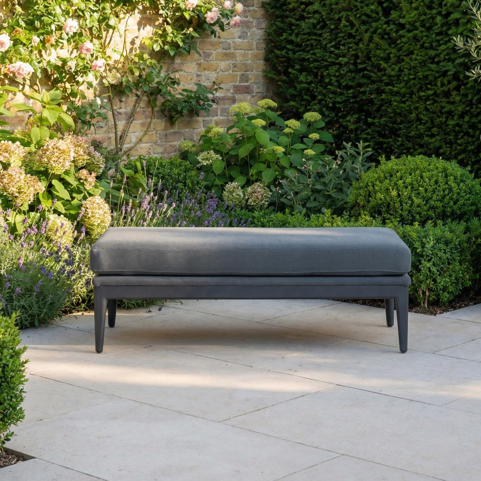 The Luna Bench in Grey is a stylish outdoor seat placed on a stone patio, surrounded by greenery and flowers—perfect as an all-weather bench for any garden retreat.