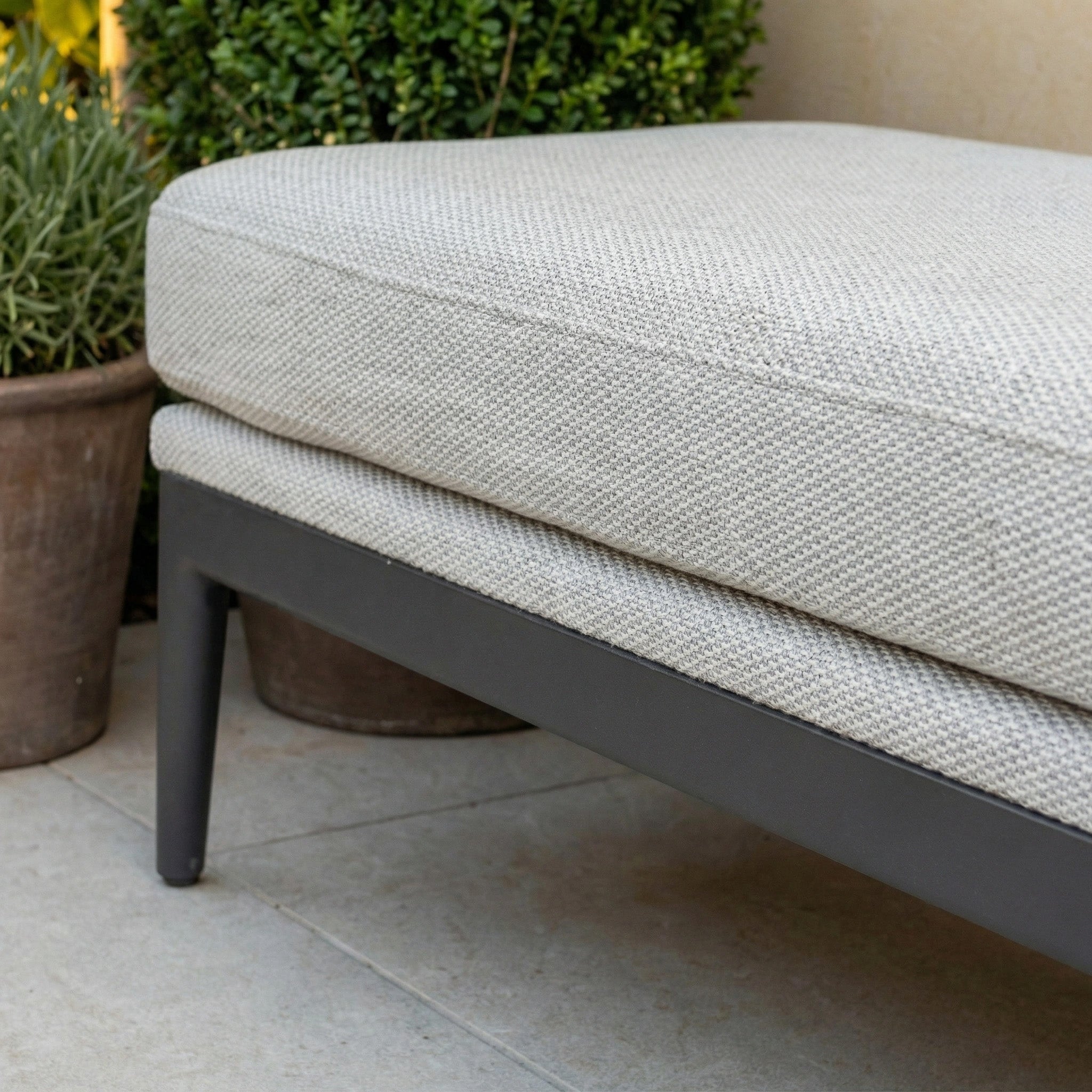 Discover the Luna Bench in Oyster Grey—an all-weather seat featuring a black frame, light grey cushion, and surrounded by potted greenery on a tiled patio. Perfect for stylish, comfortable outdoor relaxation.