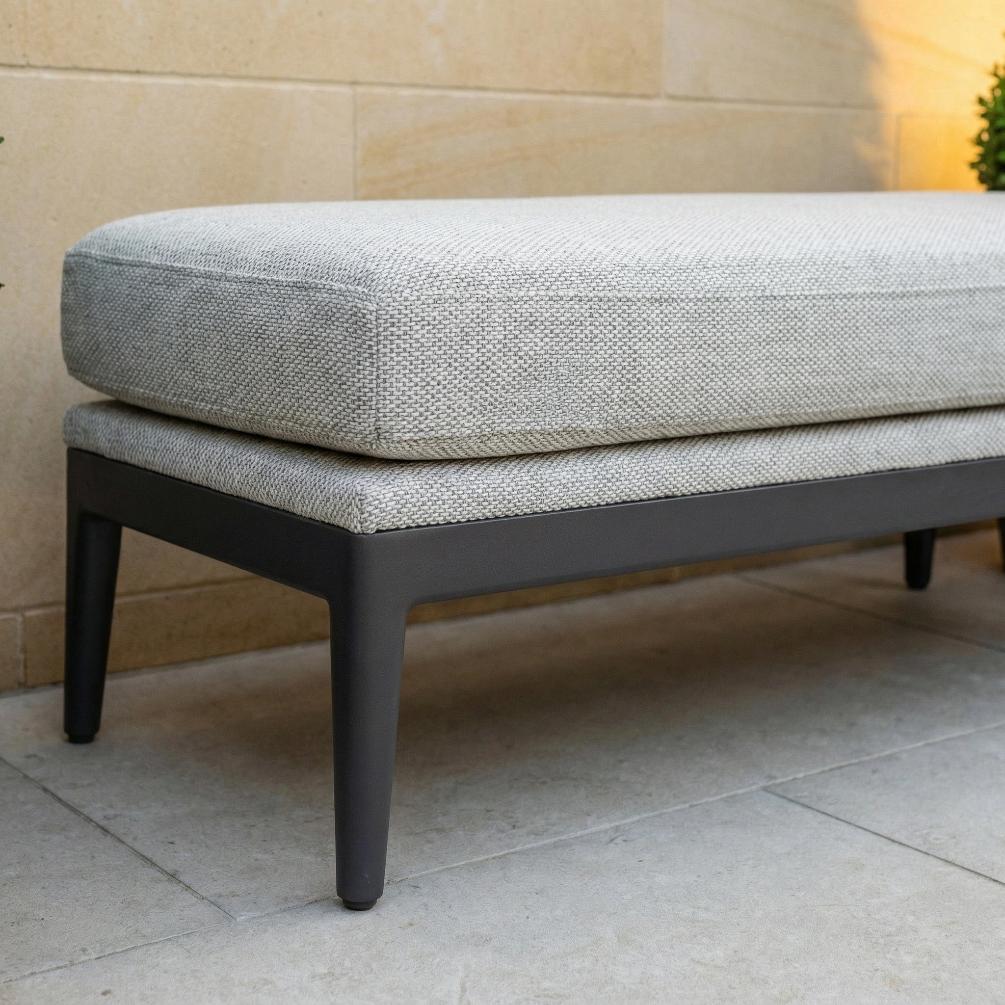 The Luna Bench in Oyster Grey features all-weather durability, a light gray cushion, and dark legs—ideal for stylish outdoor seating on a stone patio near a wall.