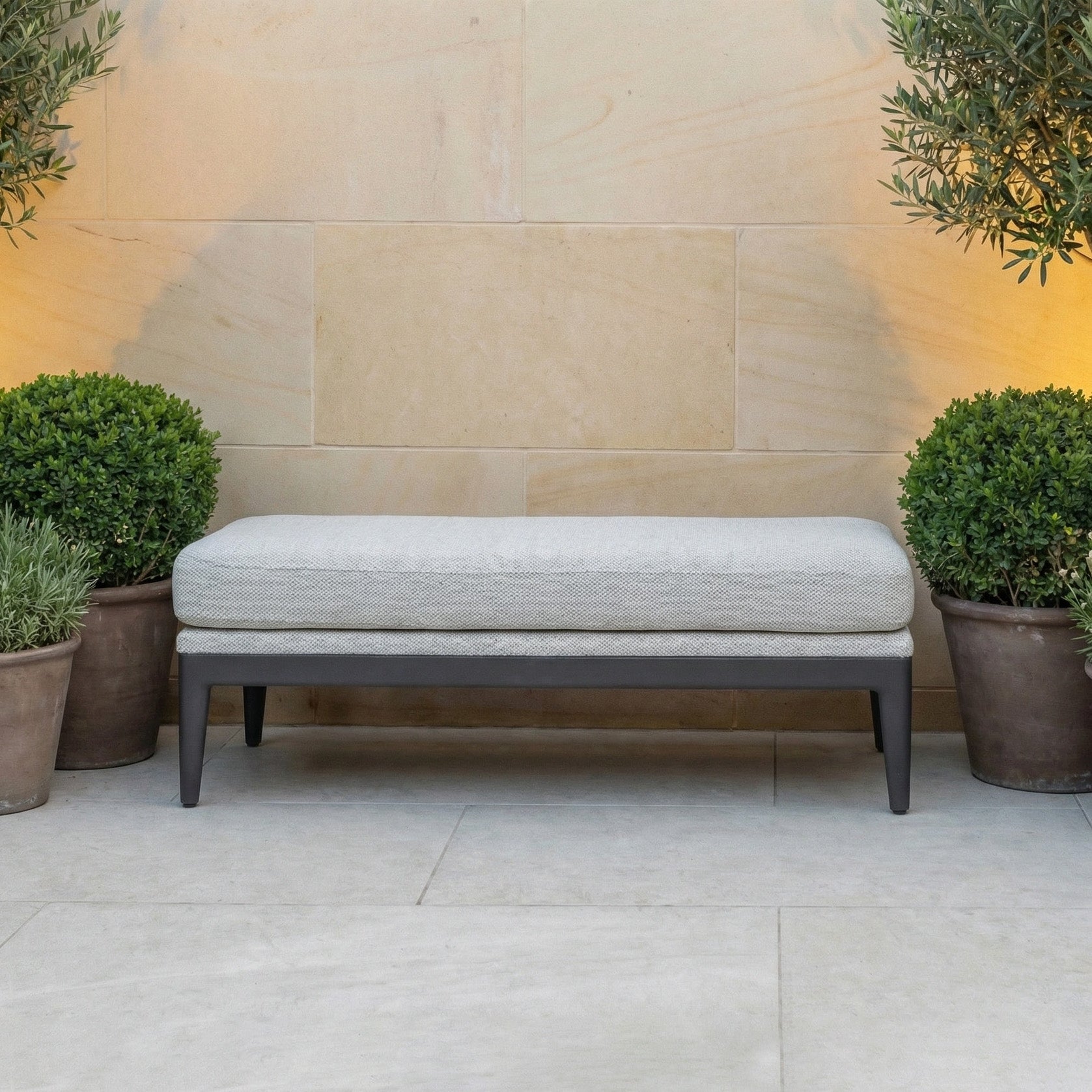 The Luna Bench in Oyster Grey is an all-weather bench with black legs, accented by green potted plants and set against a beige stone wall—ideal for stylish outdoor seating.