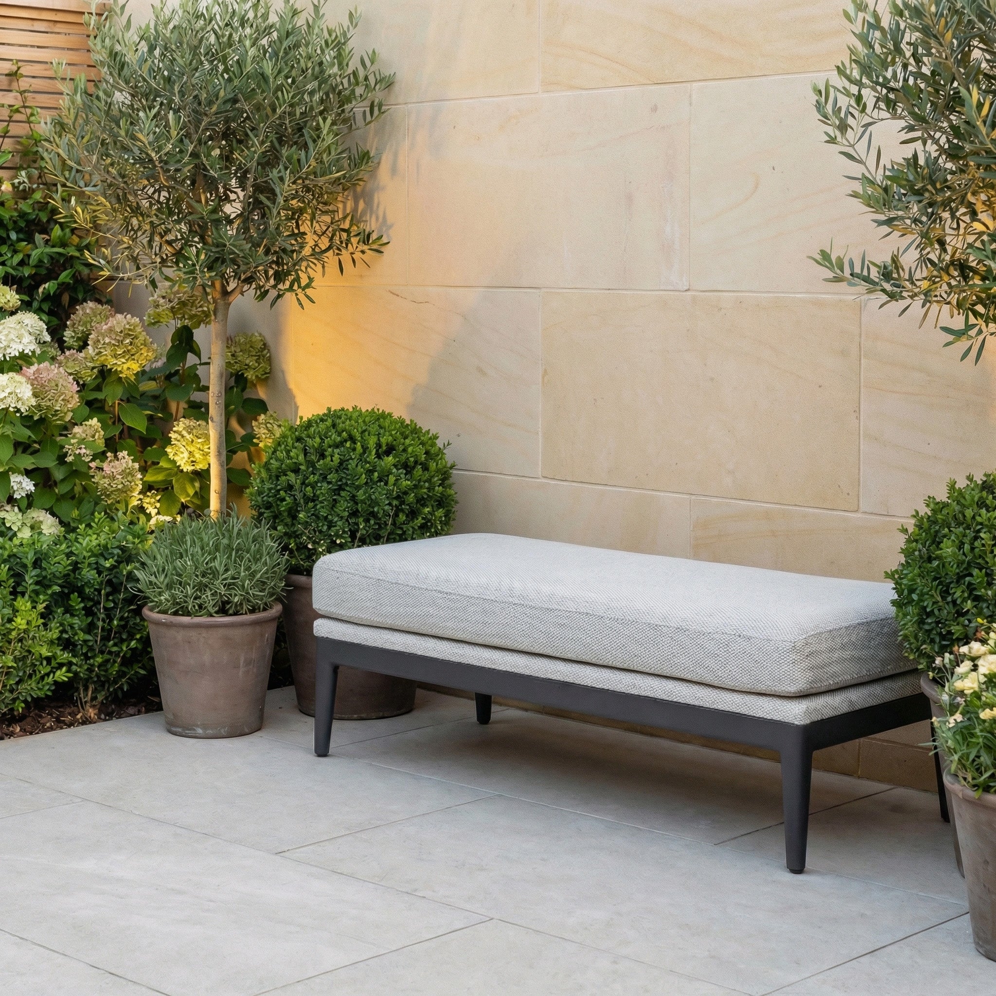 The Luna Bench in Oyster Grey with a gray cushion offers inviting, all-weather outdoor seating, perfect for placing among potted plants and greenery beside a stone wall.