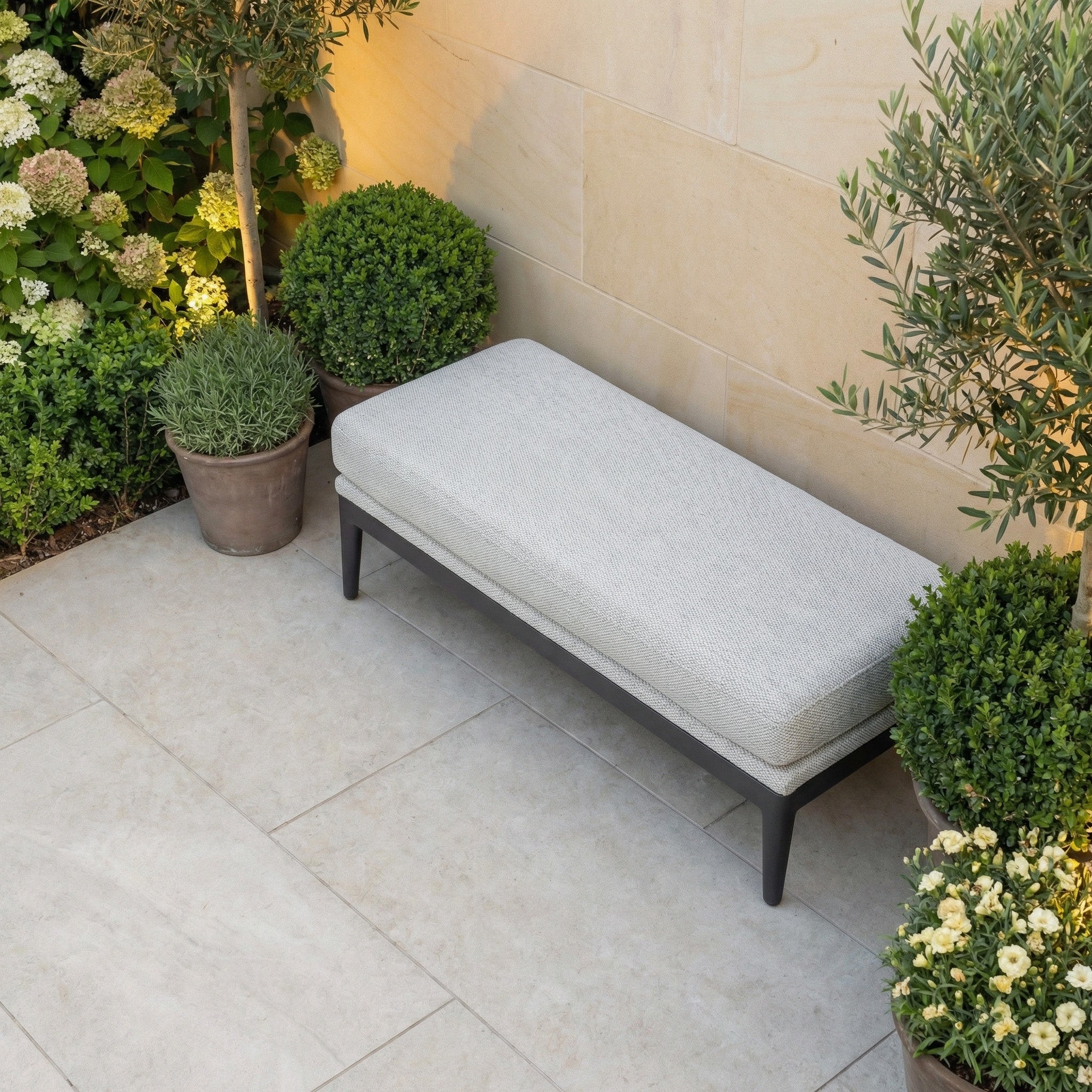 The Luna Bench in Oyster Grey, a cushioned all-weather seat, is placed on a stone patio amid potted plants and lush greenery.
