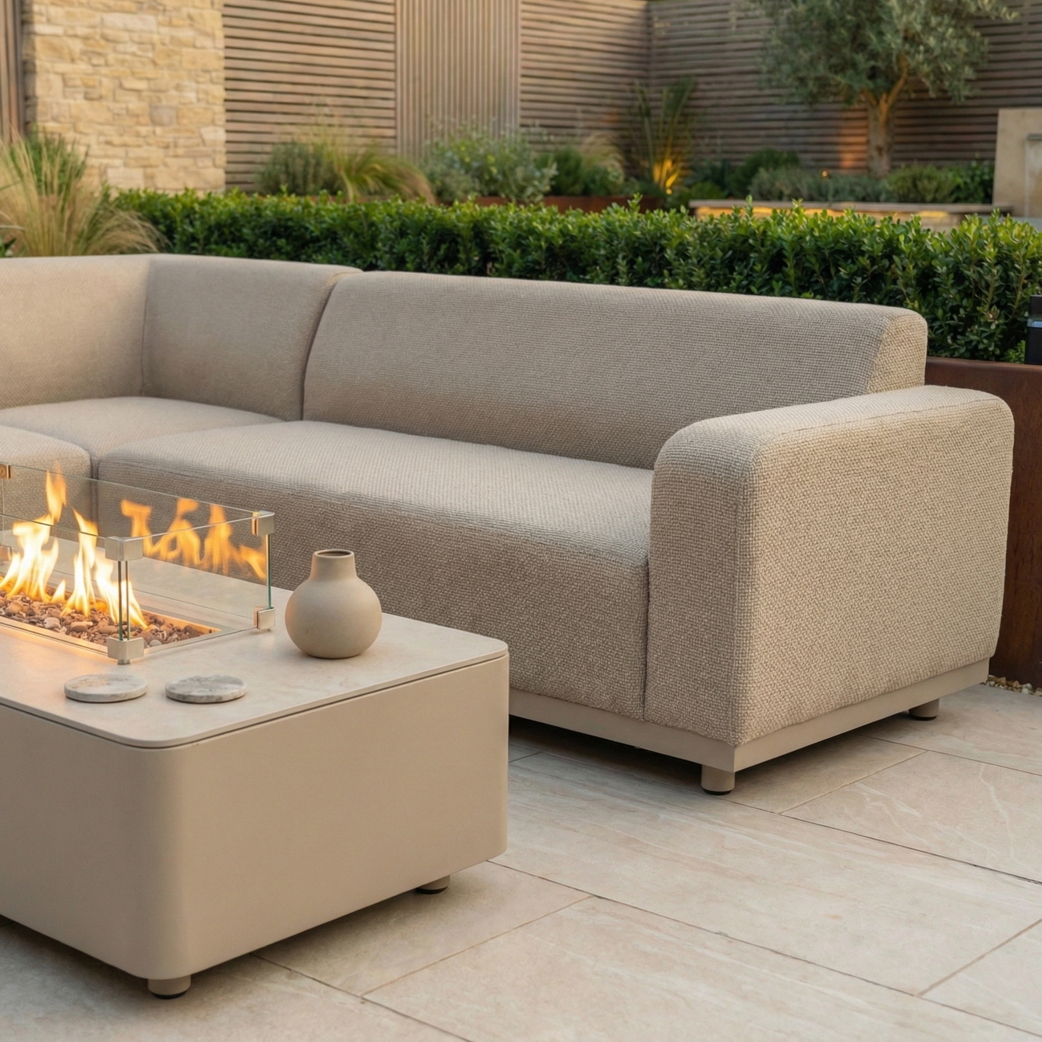 The Luna Outdoor Fabric Corner Sofa Set with Firepit Coffee Table in Fawn sits on a tiled patio, crafted from all-weather material and upholstered in Luna fabric, accented with a vase and surrounded by greenery.