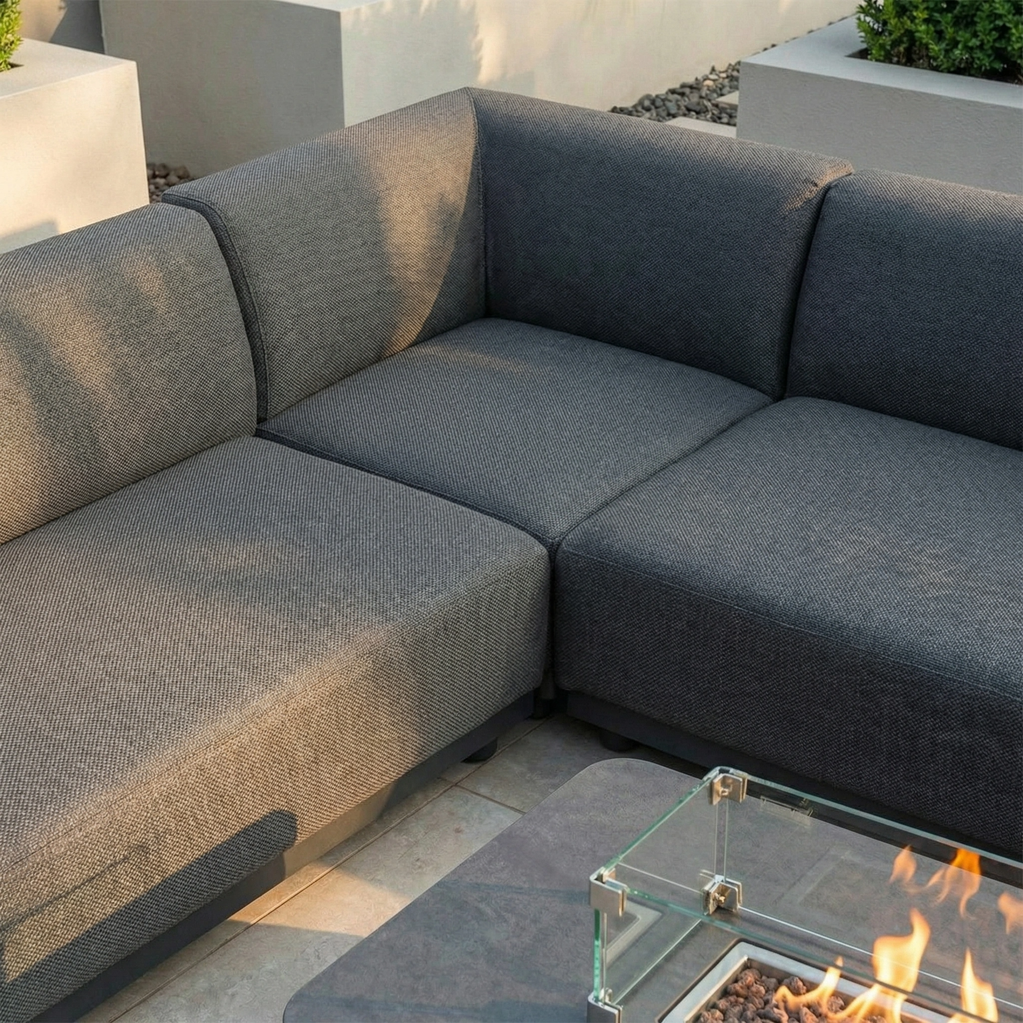 The Luna Outdoor Fabric Corner Sofa Set with Firepit Coffee Table in Grey is shown on a patio surrounded by greenery.