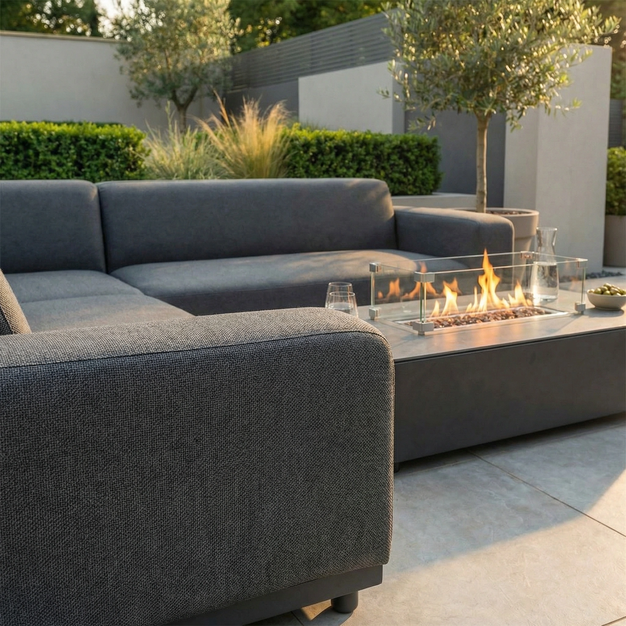 The Luna Outdoor Fabric Corner Sofa Set with Firepit Coffee Table in Grey sits on the patio, surrounded by lush green shrubs and trees.