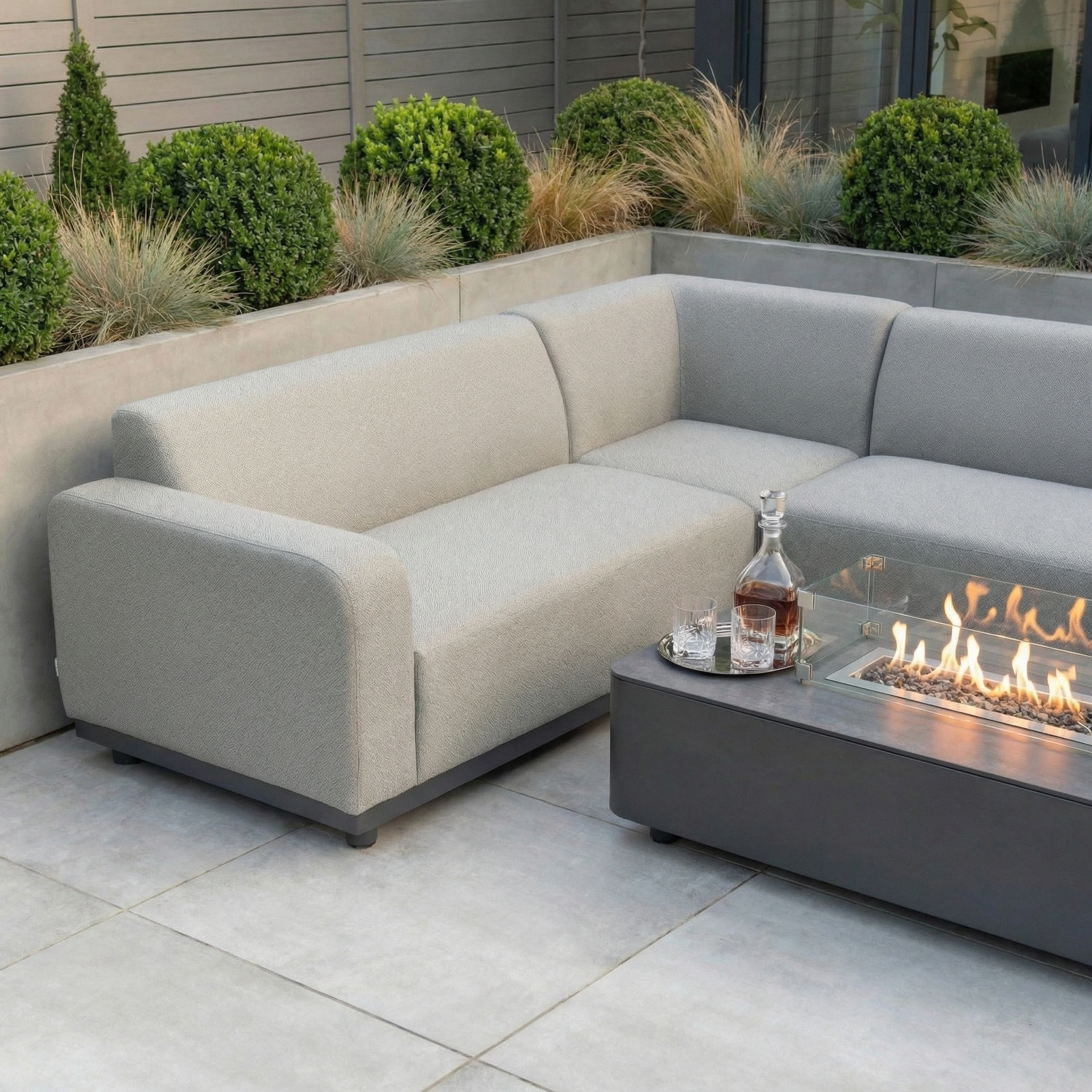 Contemporary patio setup with the Luna Outdoor Fabric Corner Sofa Set and Firepit Coffee Table in Oyster Grey, accented by glassware and a decanter.