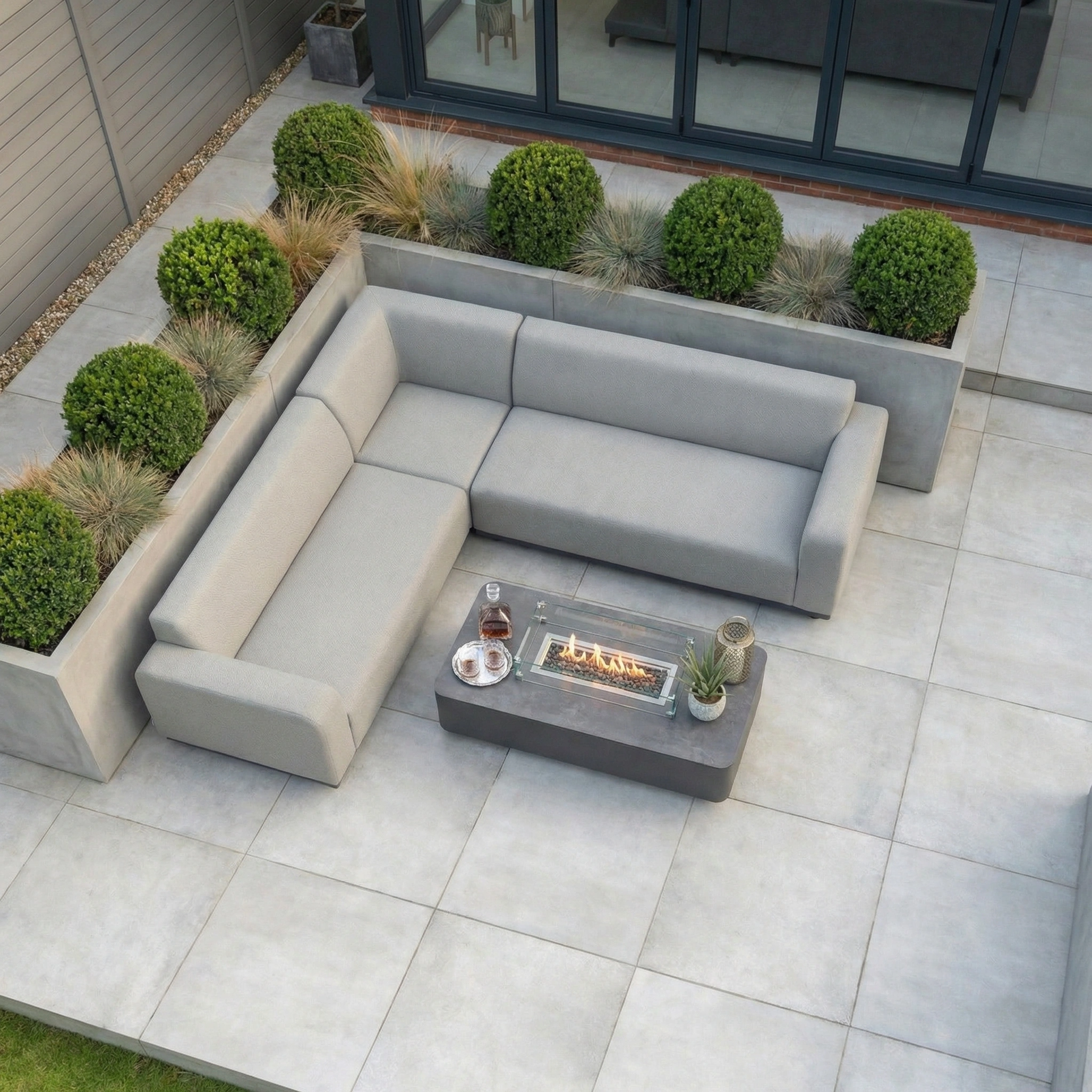 Modern patio featuring the Luna Outdoor Fabric Corner Sofa Set with Firepit Coffee Table in Oyster Grey and potted plants on a tiled floor.