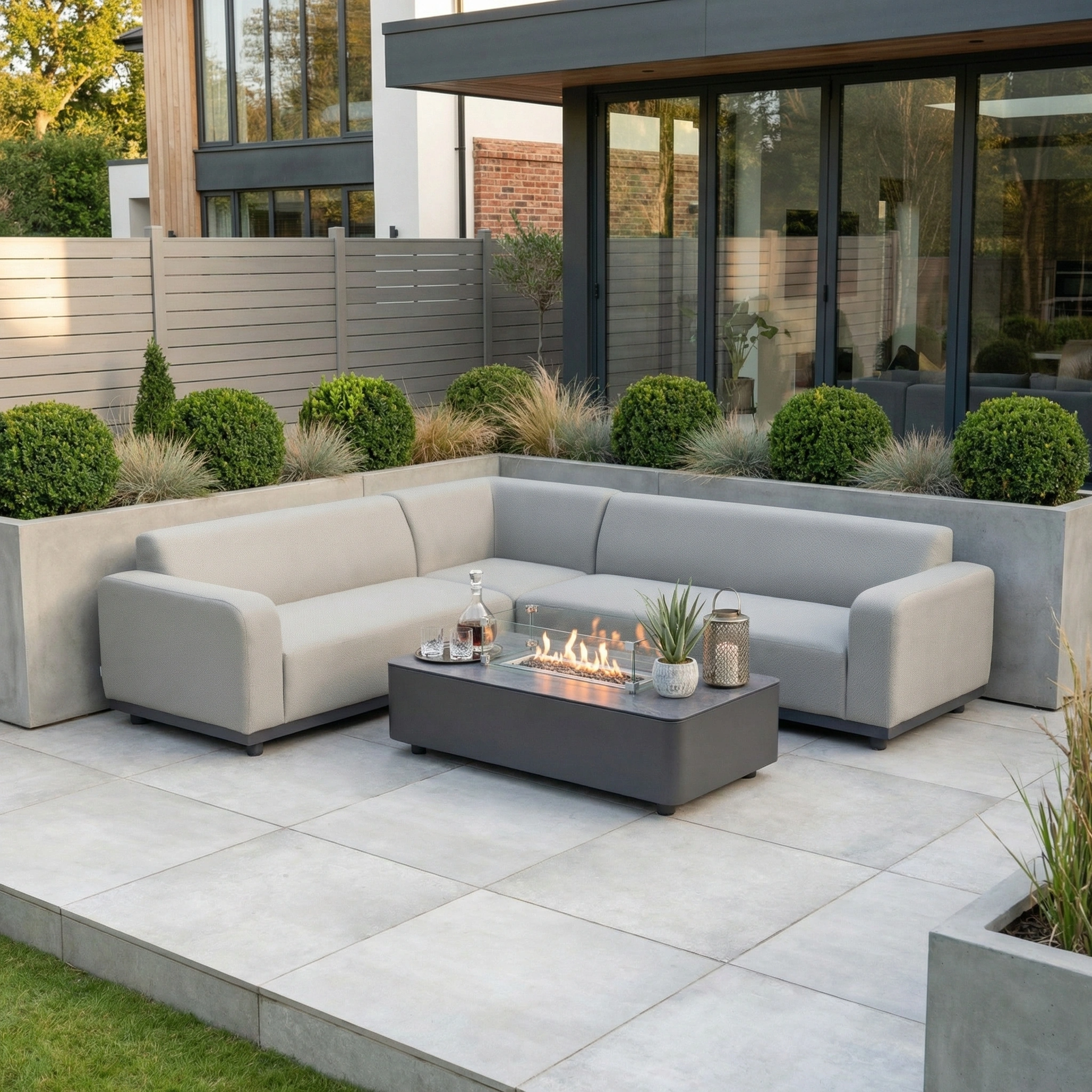 Contemporary patio featuring the Luna Outdoor Fabric Corner Sofa Set with Firepit Coffee Table in Oyster Grey, plus drinks and plants outside a modern home.