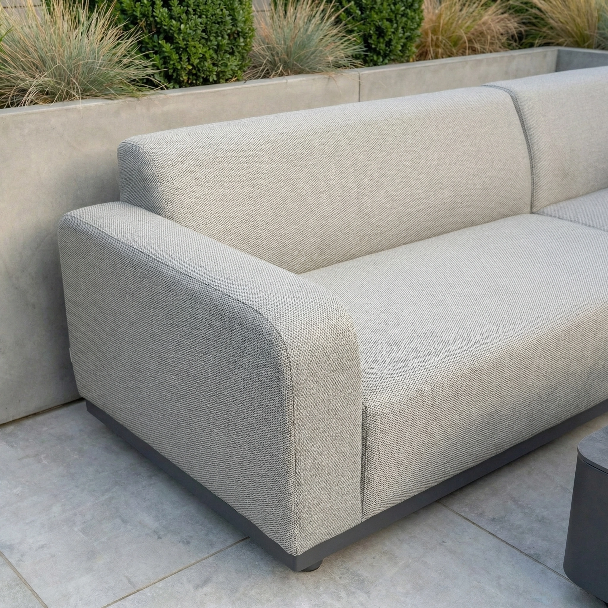 The Luna Outdoor Fabric Corner Sofa Set in Oyster Grey, paired with a firepit coffee table, sits on a tiled patio and is surrounded by lush greenery.