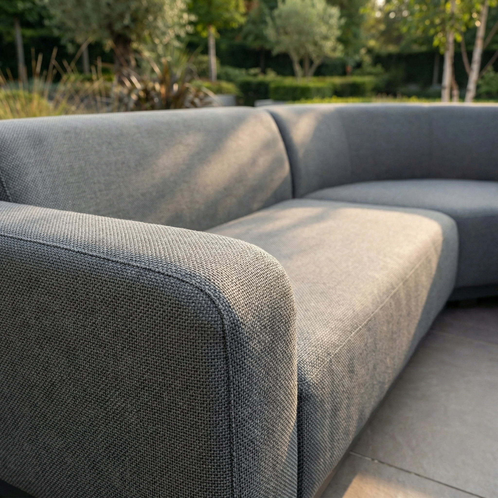 Close-up of the Luna Outdoor Fabric Curved Corner Sofa Set with Altura Rising Table in Grey, displayed on a patio with greenery in the background.