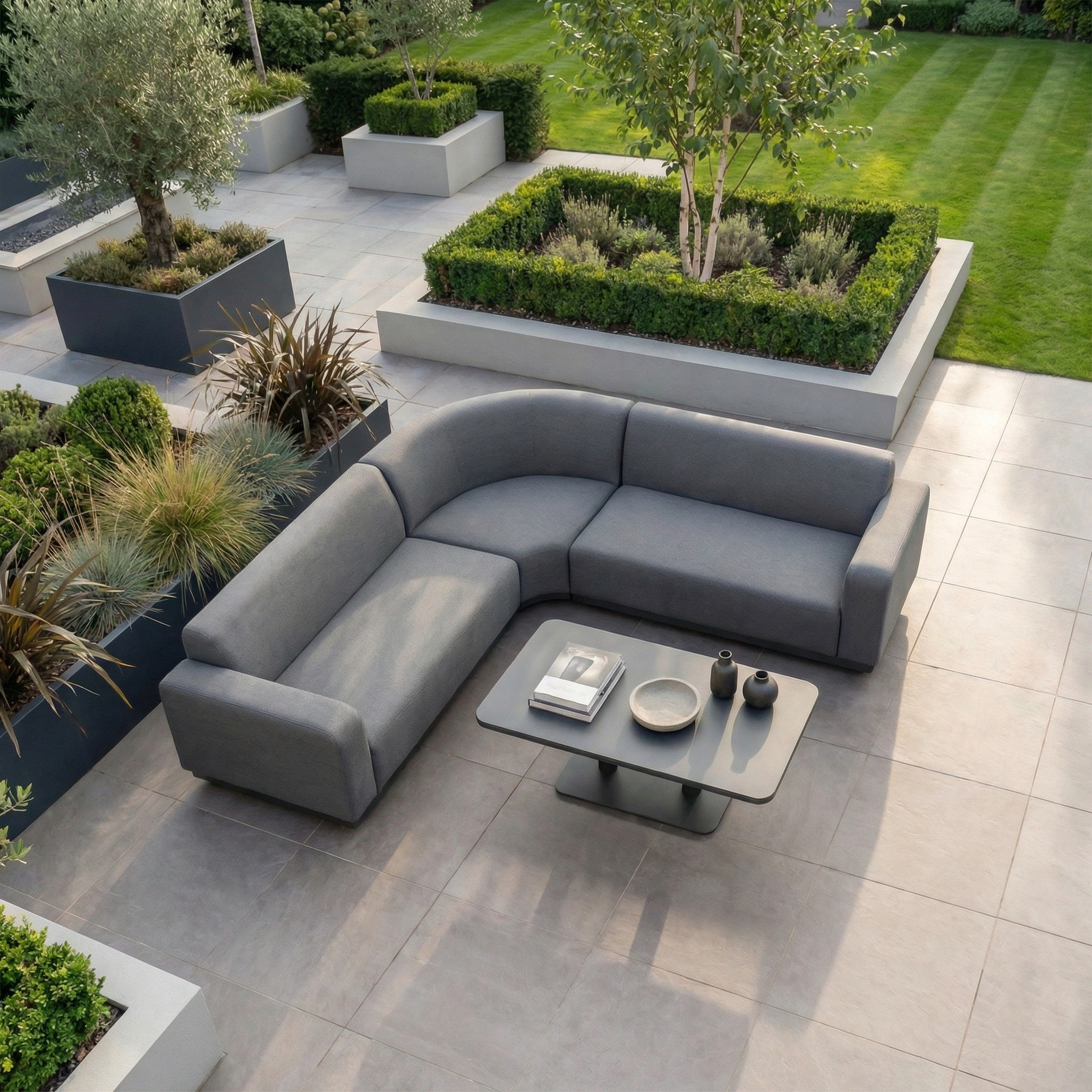 Modern outdoor patio featuring the Luna Outdoor Fabric Curved Corner Sofa Set with Altura Rising Table in Grey, surrounded by landscaped garden beds and tiled flooring.