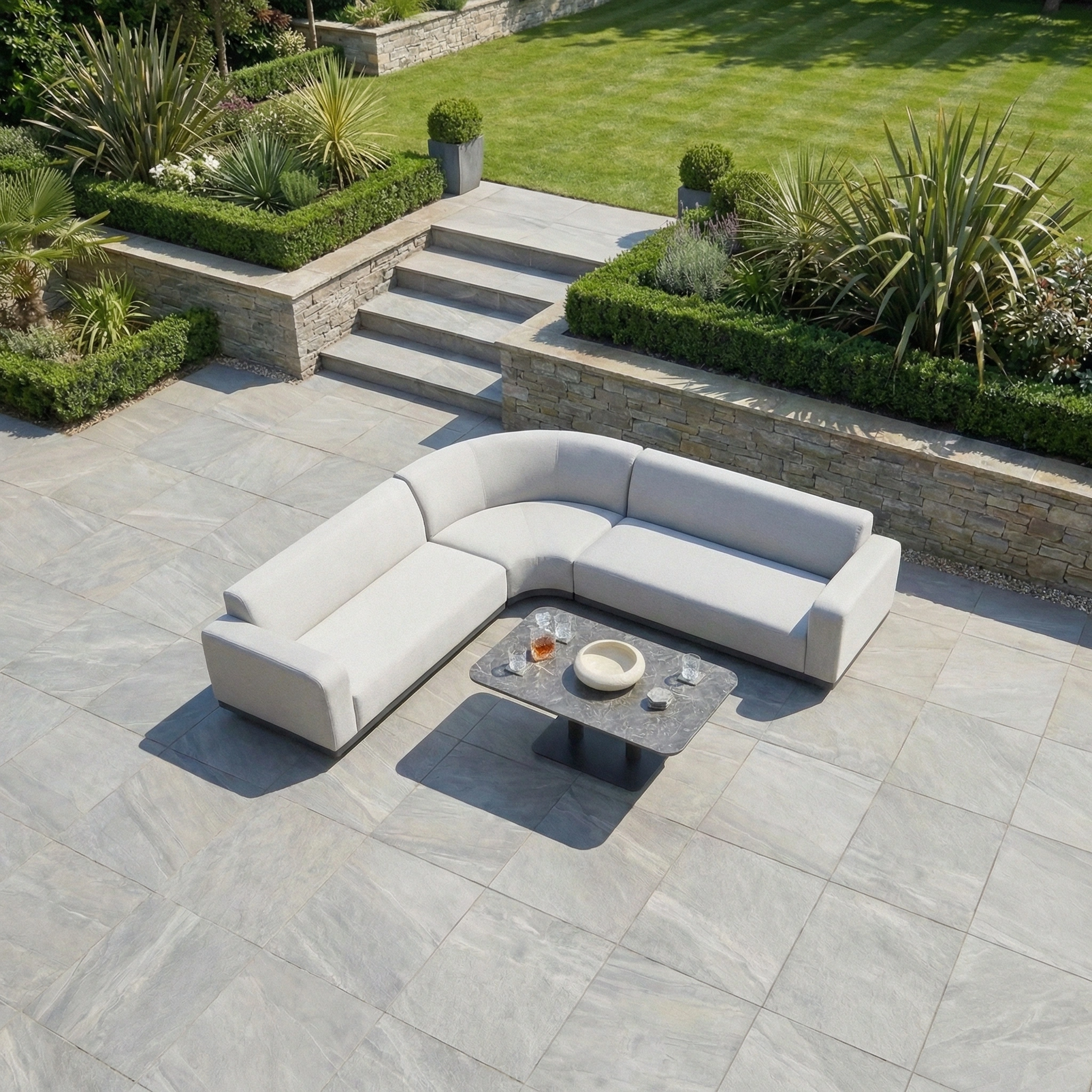 A modern patio with the Luna Outdoor Fabric Curved Corner Sofa Set and Altura Rising Table in Oyster Grey sits on stone tiles, showcasing all-weather furniture near a landscaped garden.