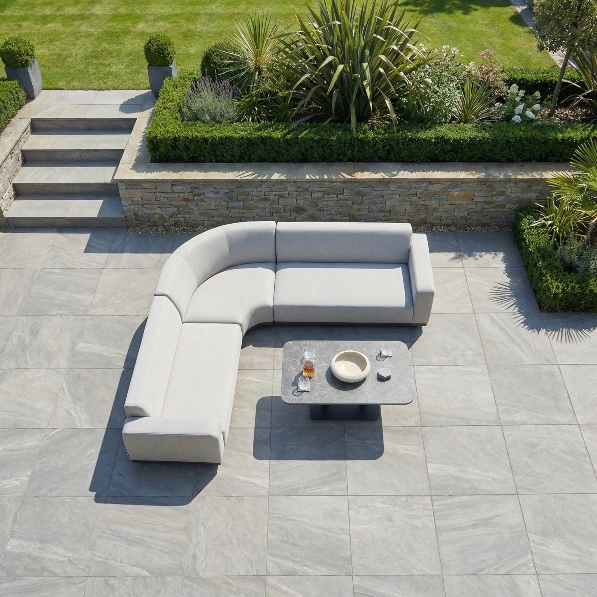 The Luna Outdoor Fabric Curved Corner Sofa Set with Altura Rising Table in Oyster Grey sits on a tiled patio, offering stylish all-weather comfort, with a landscaped garden and steps providing a scenic backdrop.