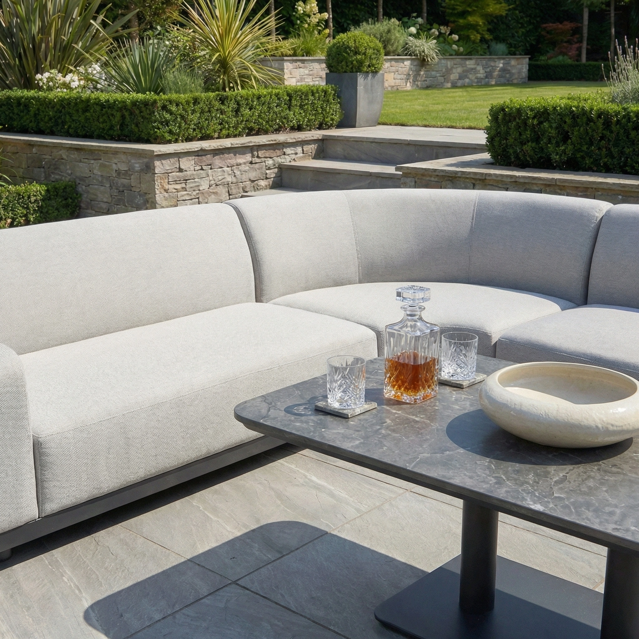 The Luna Outdoor Fabric Curved Corner Sofa Set with Altura Rising Table in Oyster Grey offers all-weather comfort, stylishly arranged in a landscaped garden with drinks and a bowl on its stone-effect rising table.