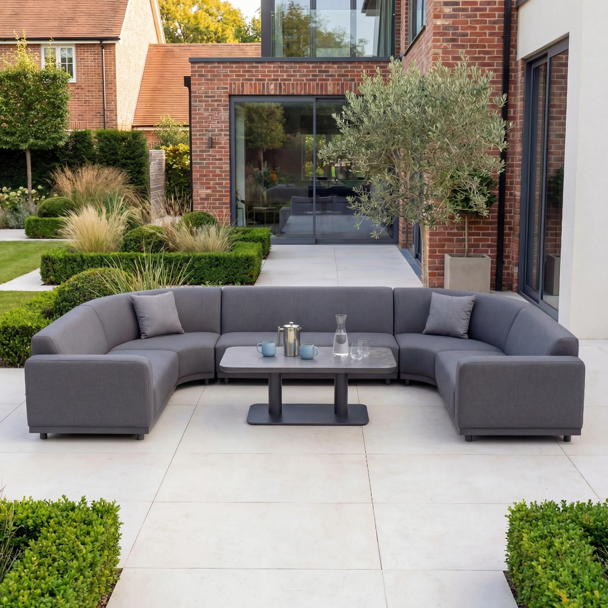 Modern outdoor patio featuring the Luna Outdoor Fabric Curved U-Shape Sofa Set with Altura Rising Table in Grey, surrounded by lush greenery in a landscaped garden.