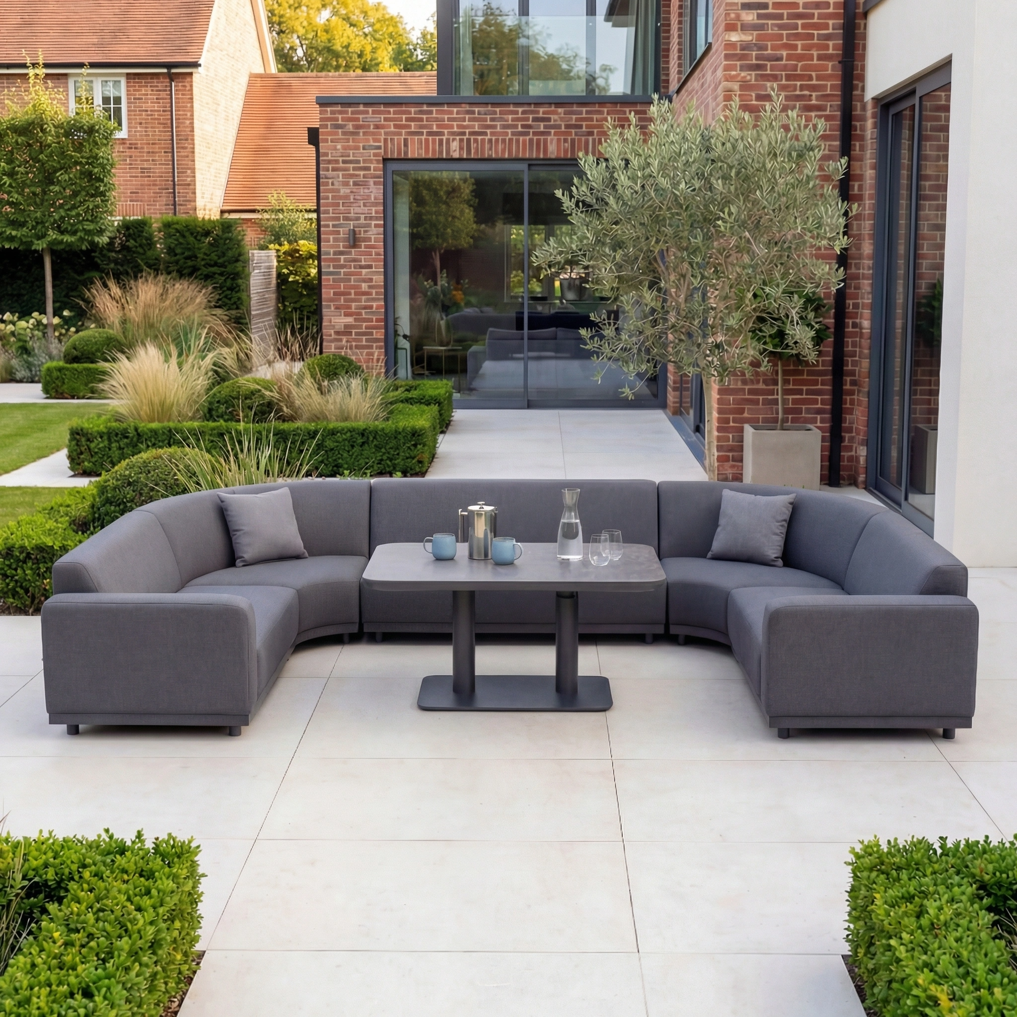 The Luna Outdoor Fabric Curved U-Shape Sofa Set with Altura Rising Table in Grey is stylish all-weather garden furniture, ideal for relaxing outdoors on a modern patio surrounded by plants and a brick house.