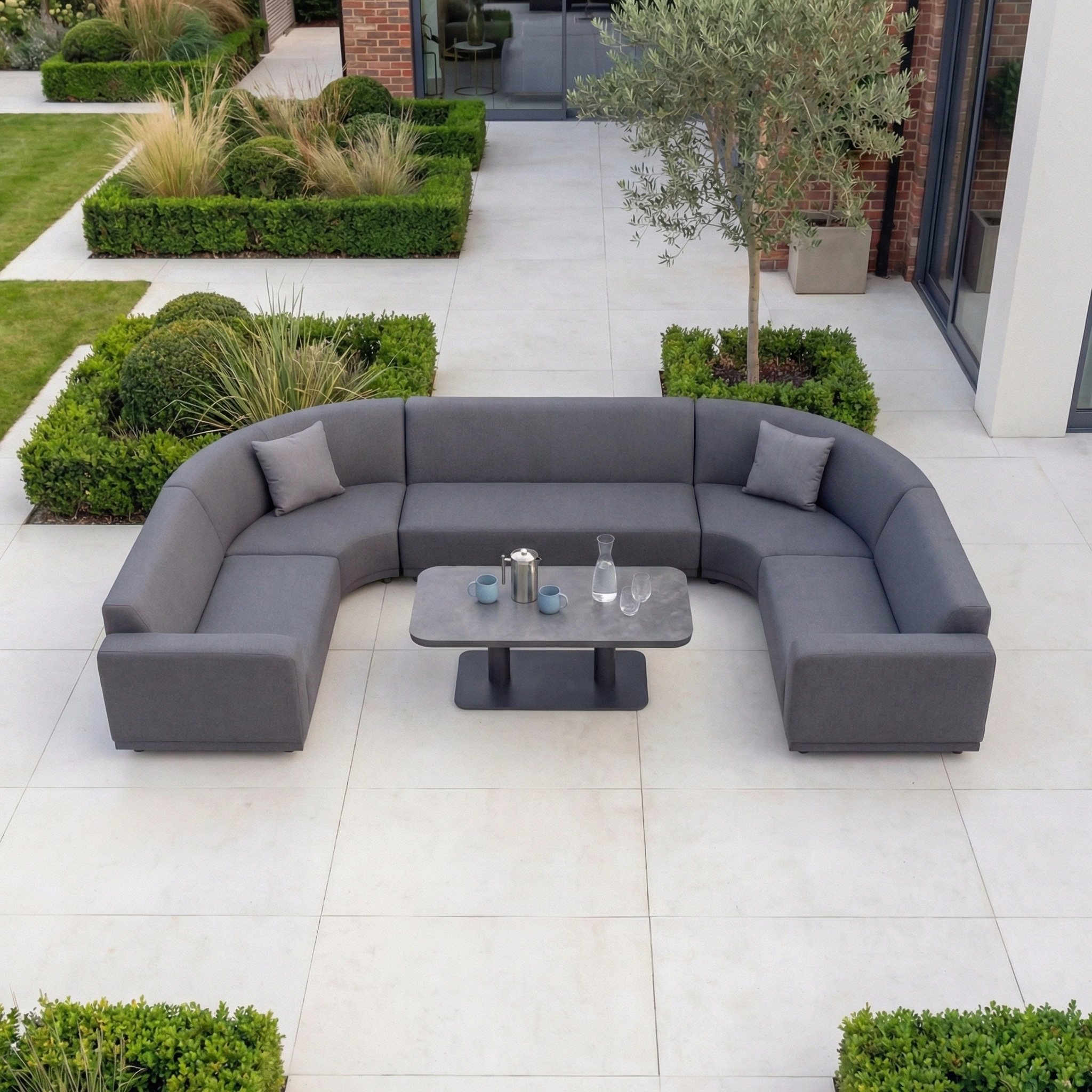 The Luna Outdoor Fabric Curved U-Shape Sofa Set with Altura Rising Table in Grey features two cushions and a versatile table—stylish all-weather garden furniture perfect for any patio, surrounded by lush greenery.