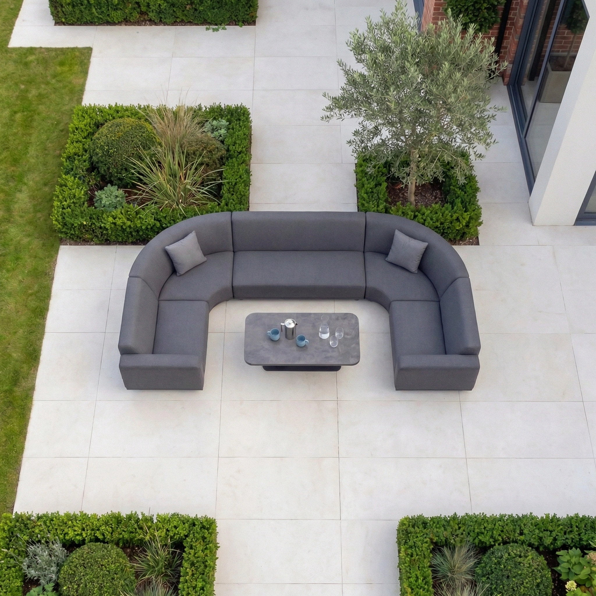 The Luna Outdoor Fabric Curved U-Shape Sofa Set with Altura Rising Table in Grey offers stylish, all-weather comfort with plush pillows—a perfect centerpiece for relaxing outdoors on your patio amid lush greenery.