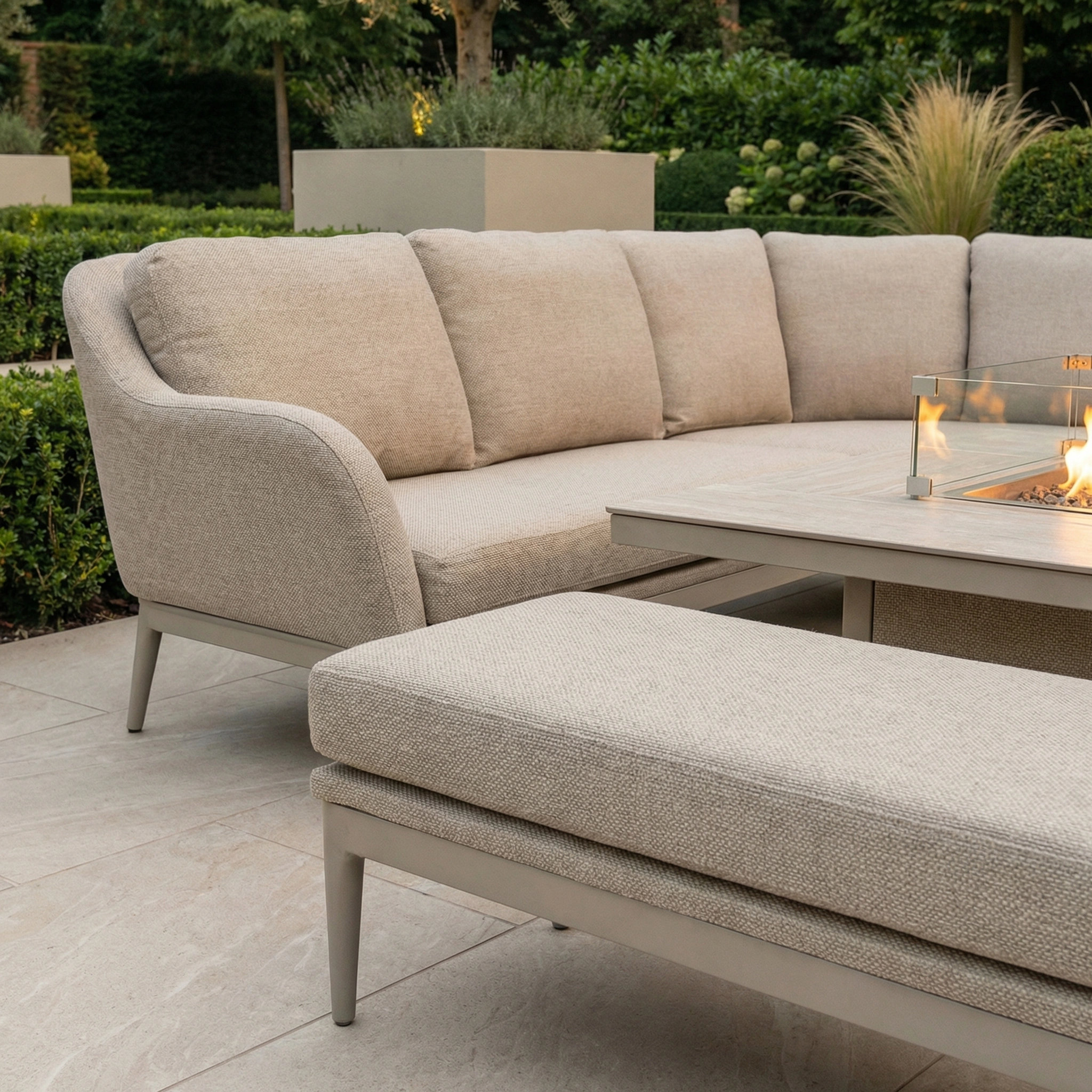 The Luna Deluxe Outdoor Fabric Square Corner Dining Set with Rising Firepit Table in Fawn sits on a stone patio in a garden setting.