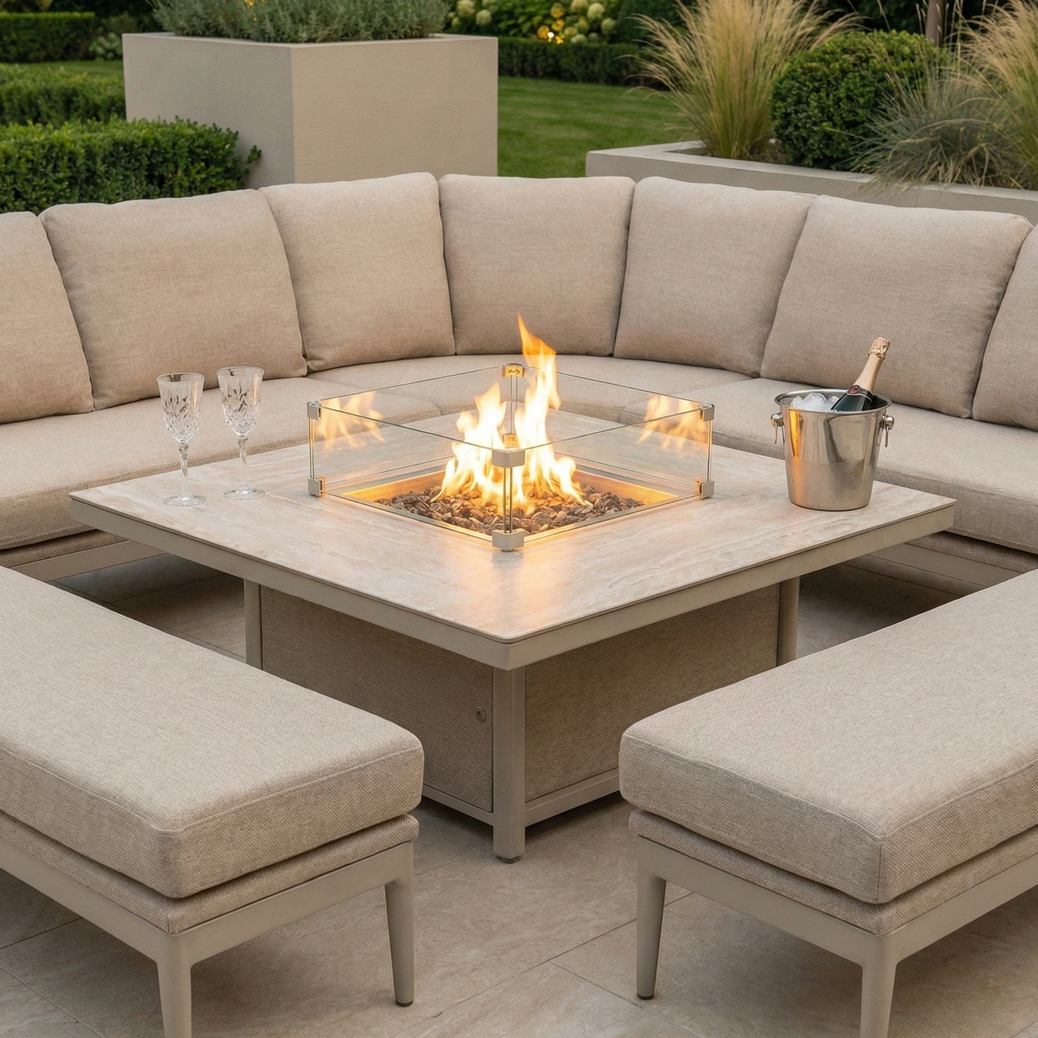 A cozy outdoor seating area with the Luna Deluxe Outdoor Fabric Square Corner Dining Set in Fawn, featuring a rising firepit table, champagne, and glasses.