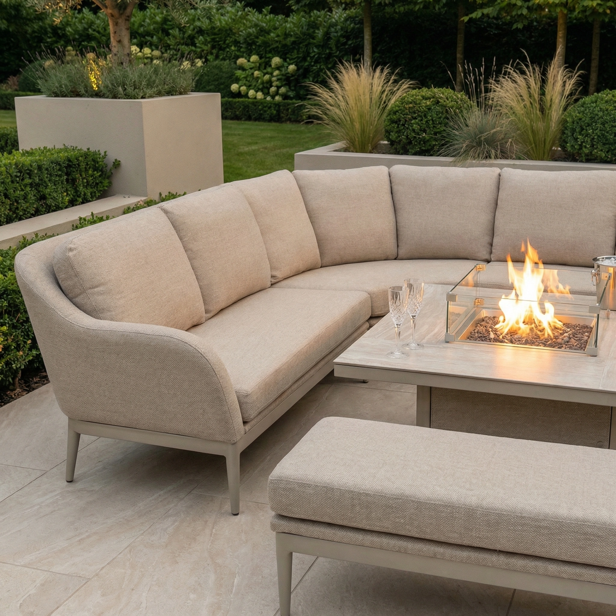 Luna Deluxe Outdoor Fabric Square Corner Dining Set with Rising Firepit Table in Fawn on a patio, surrounded by greenery and decorative plants.