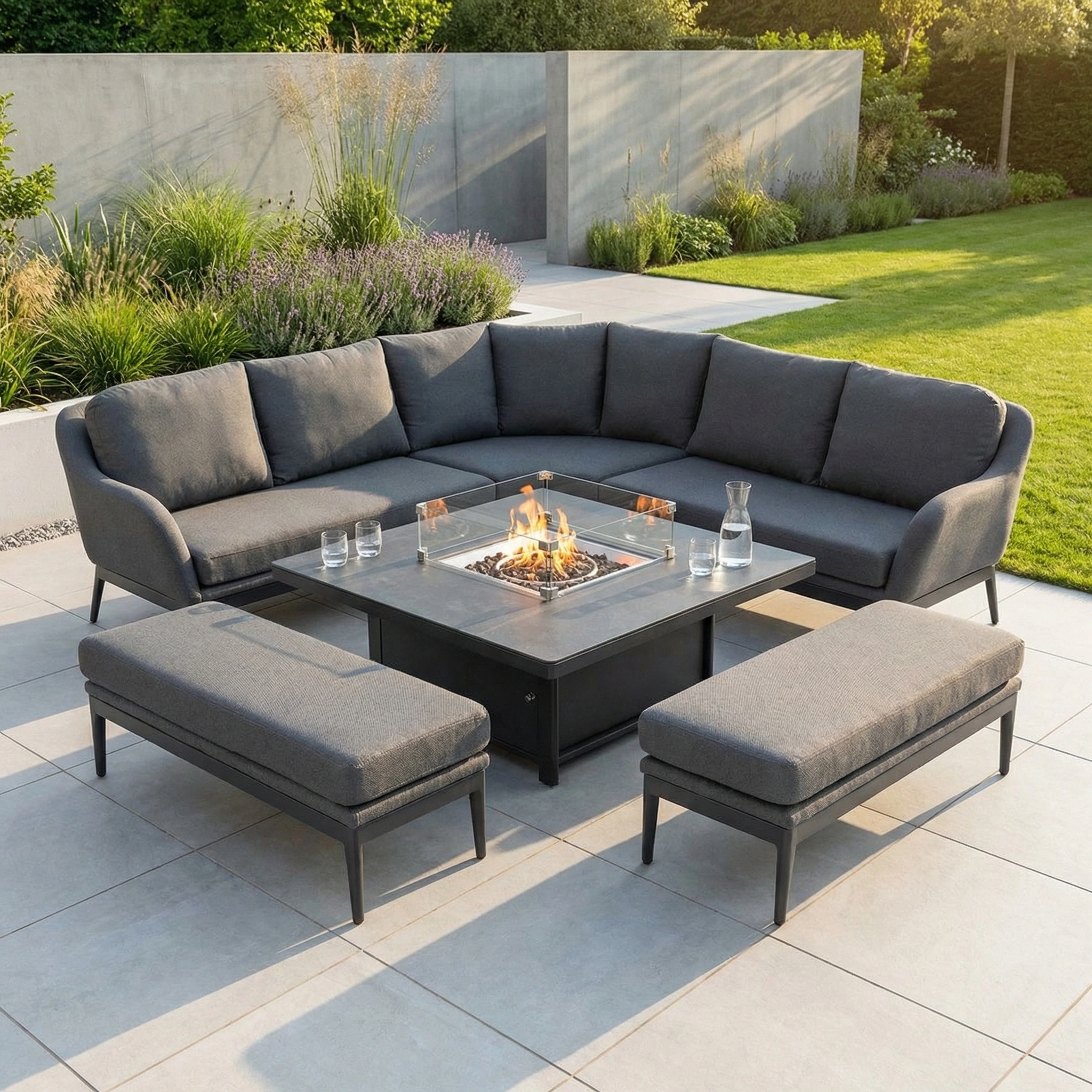 The Luna Deluxe Outdoor Fabric Square Corner Dining Set with Rising Firepit Table in Grey is perfect for relaxing or entertaining on your patio, featuring benches and weather-resistant style surrounded by greenery.