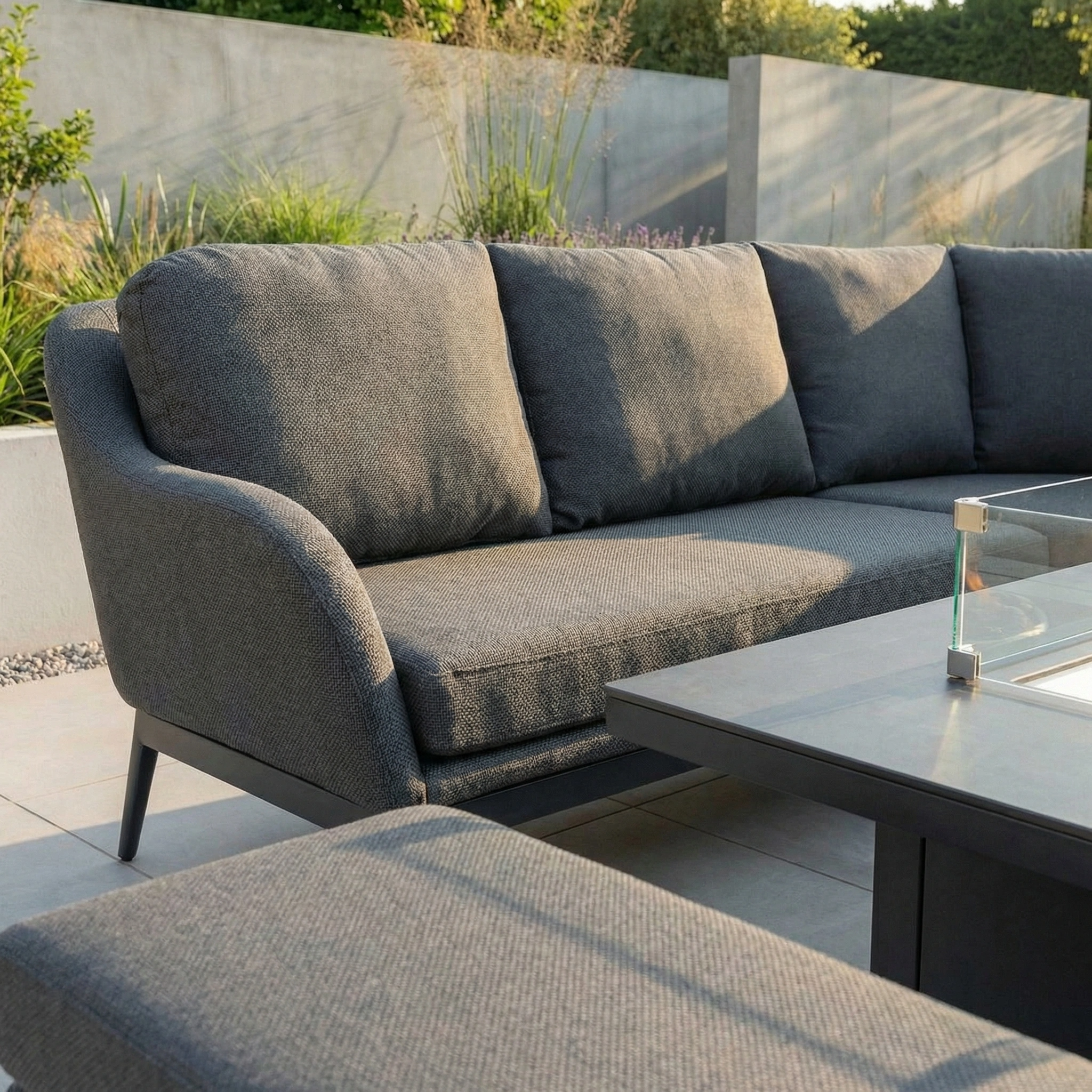The Luna Deluxe Outdoor Fabric Square Corner Dining Set with Rising Firepit Table in Grey sits on a tiled patio, offering stylish all-weather comfort with plush cushions and lush garden greenery in the background.
