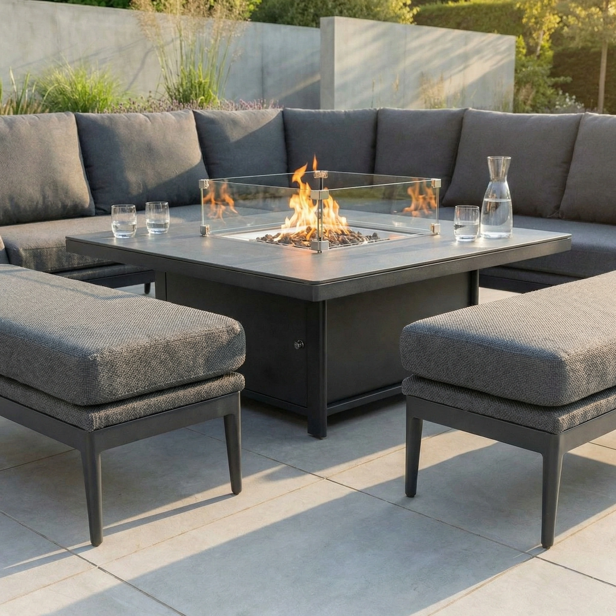 Modern patio featuring the Luna Deluxe Outdoor Fabric Square Corner Dining Set with Rising Firepit Table in Grey—complete with water carafe and glasses on tiled flooring, offering stylish comfort and durable all-weather design.