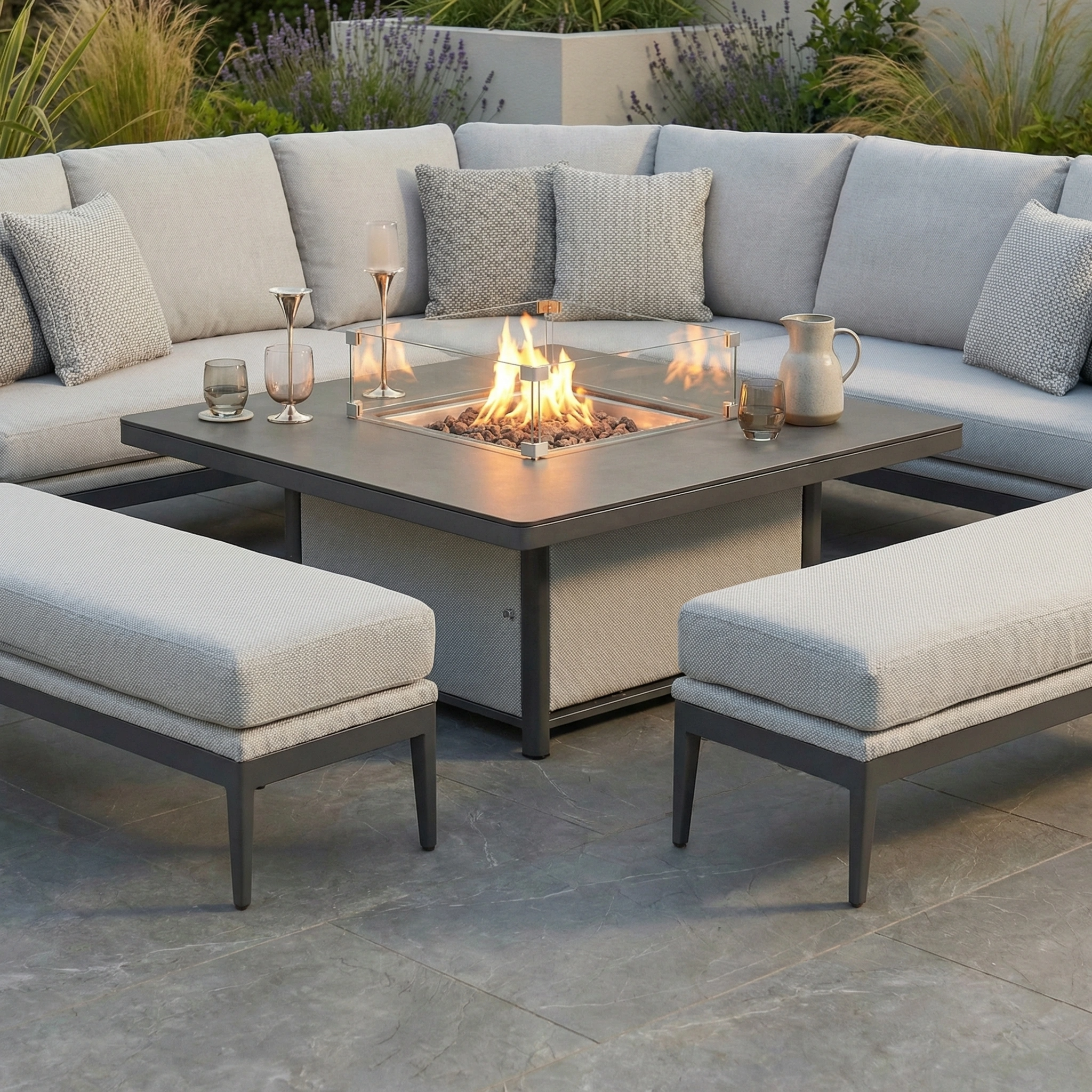 The Luna Deluxe Outdoor Fabric Square Corner Dining Set with Rising Firepit Table in Oyster Grey offers modern seating with a curved sofa, drinks beside the firepit table, cozy pillows, and greenery for a stylish outdoor retreat.