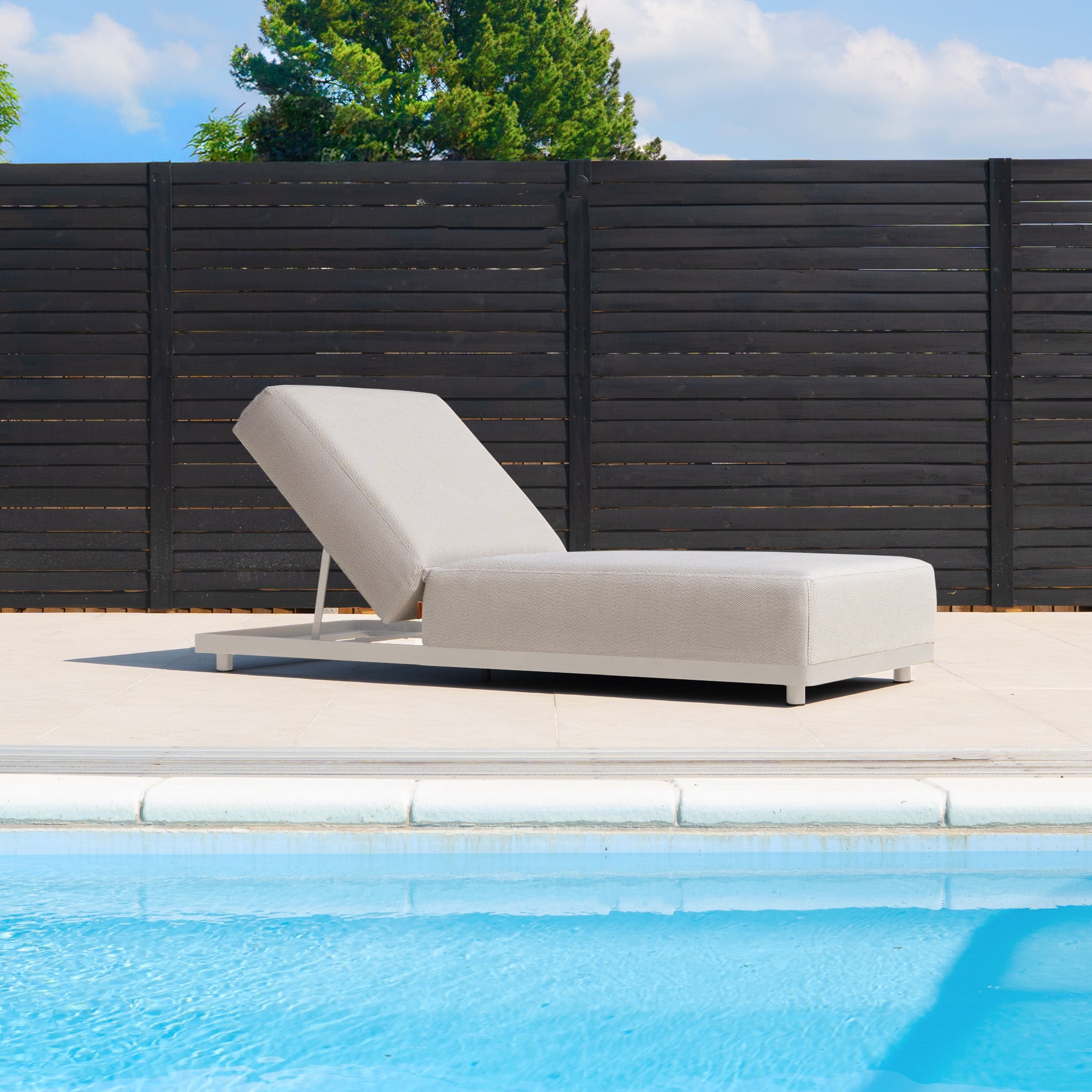 A Luna Outdoor Fabric Deluxe Sun Lounger with Bolster Cushion in Fawn sits beside a swimming pool, framed by a black fence and trees in the background.